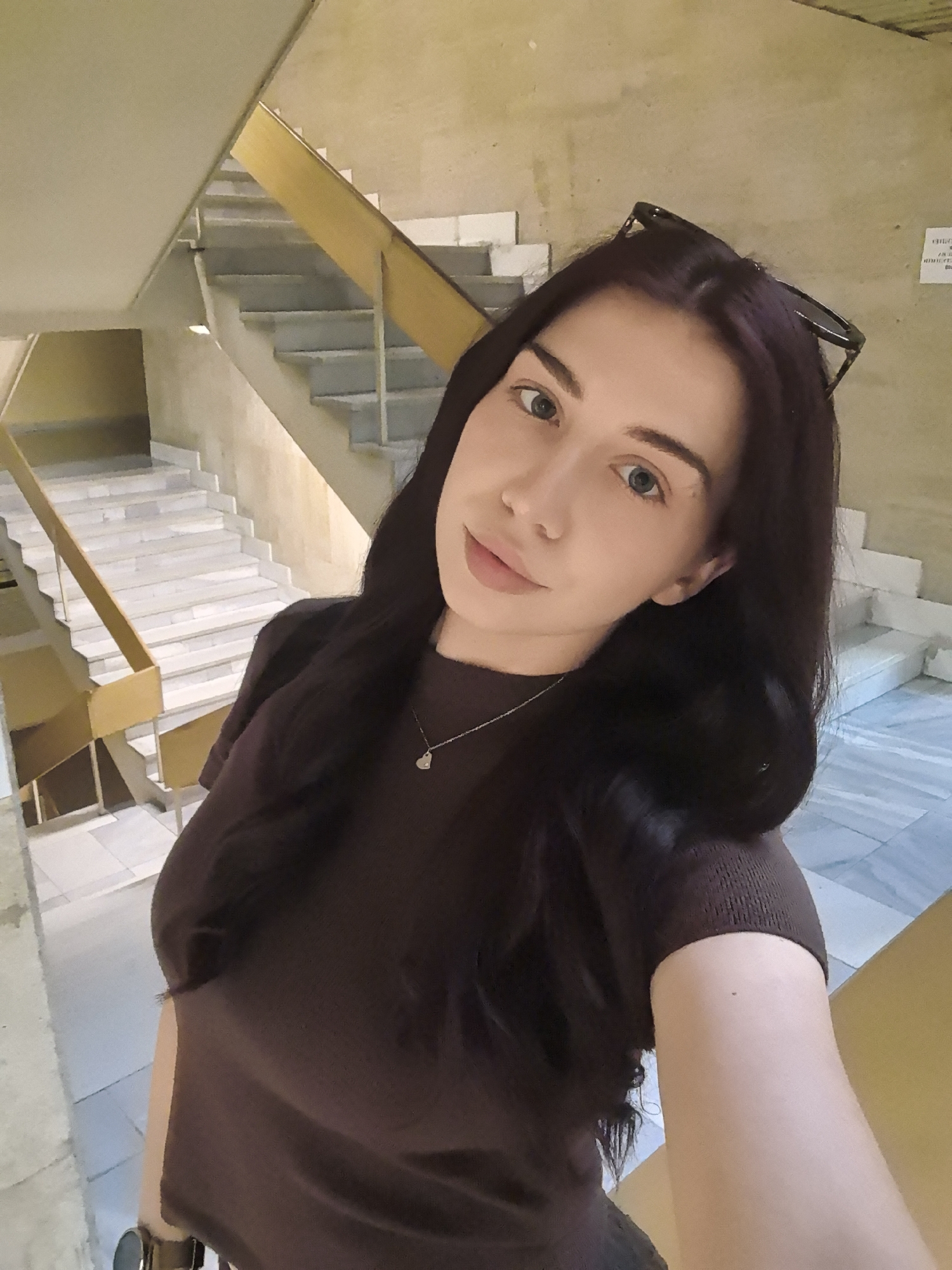 A selfie of a woman with long, dark hair, wearing a brown shirt and a silver necklace with a heart pendant. Sunglasses are perched on her head. The background shows a stairway with a yellow railing and marble-patterned flooring, suggesting an indoor public space. The location is Unknown, Unknown.