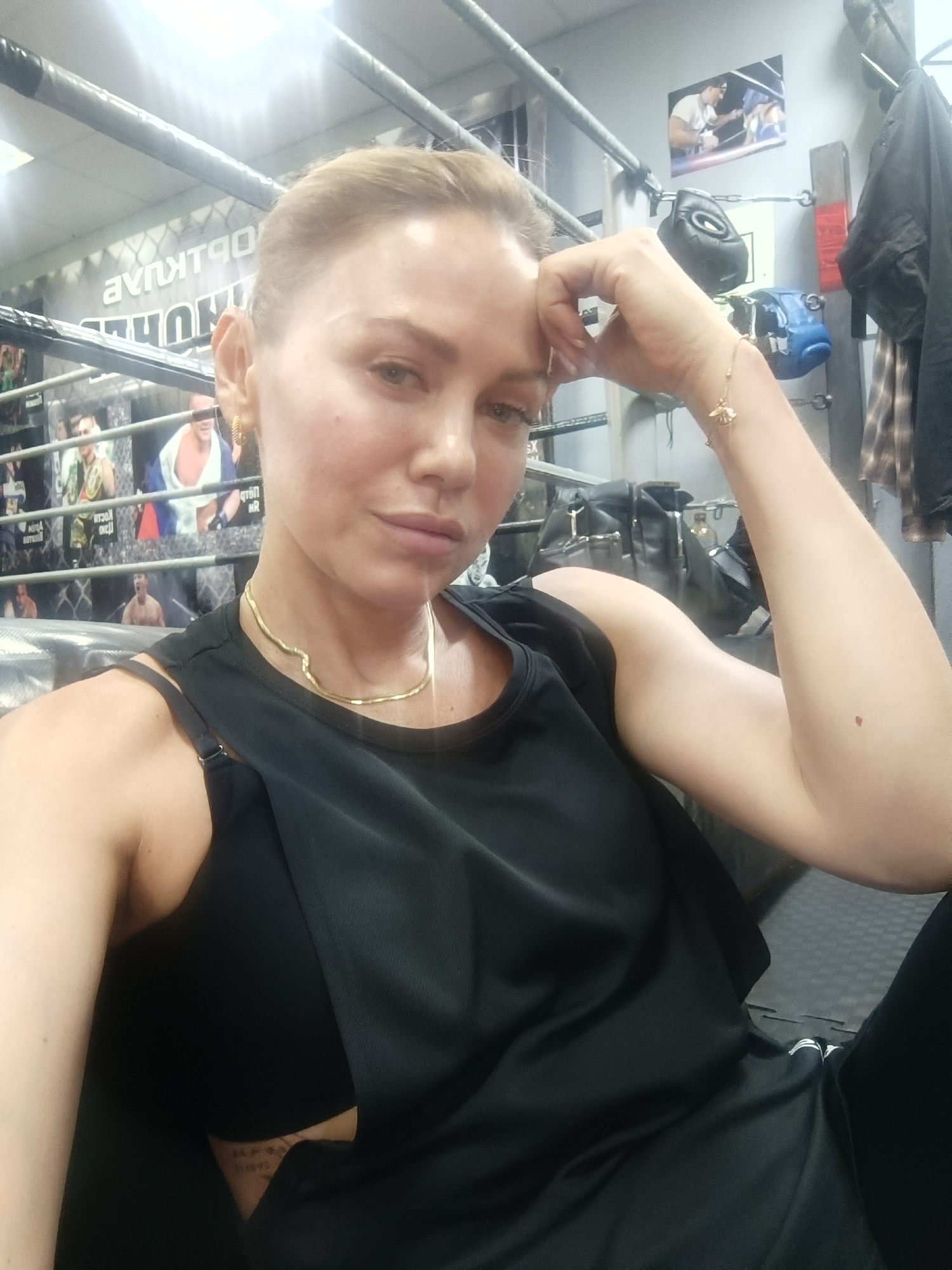 The image is a medium close-up selfie of a woman taken indoors, likely within a boxing gym or martial arts facility in Naberezhnye Chelny, Russia. The woman, with light-colored hair pulled back, looks directly at the camera with a neutral or slightly pensive expression. She wears a black sleeveless top, a gold chain necklace, and a gold bracelet featuring a bee charm on her right wrist. Her right hand rests on her forehead. A subtle, illegible tattoo is visible on her left side, partially obscured by her clothing.
The setting is clearly a gym, indicated by the background elements. The woman appears to be seated or leaning near a boxing ring, with its ropes and corner padding visible. On the walls behind her, various posters of fighters or banners are displayed. Partially visible and mirrored text on one banner reads "AVNKTPQ" and "THUHH", possibly a stylized or reversed Cyrillic gym name or slogan. To the right, boxing equipment such as black and blue punching bags, boxing gloves, and a speed bag are hung. A black backpack is also seen in the background. The space is illuminated by bright artificial lighting. The scene suggests a momentary pause or rest during a training session.