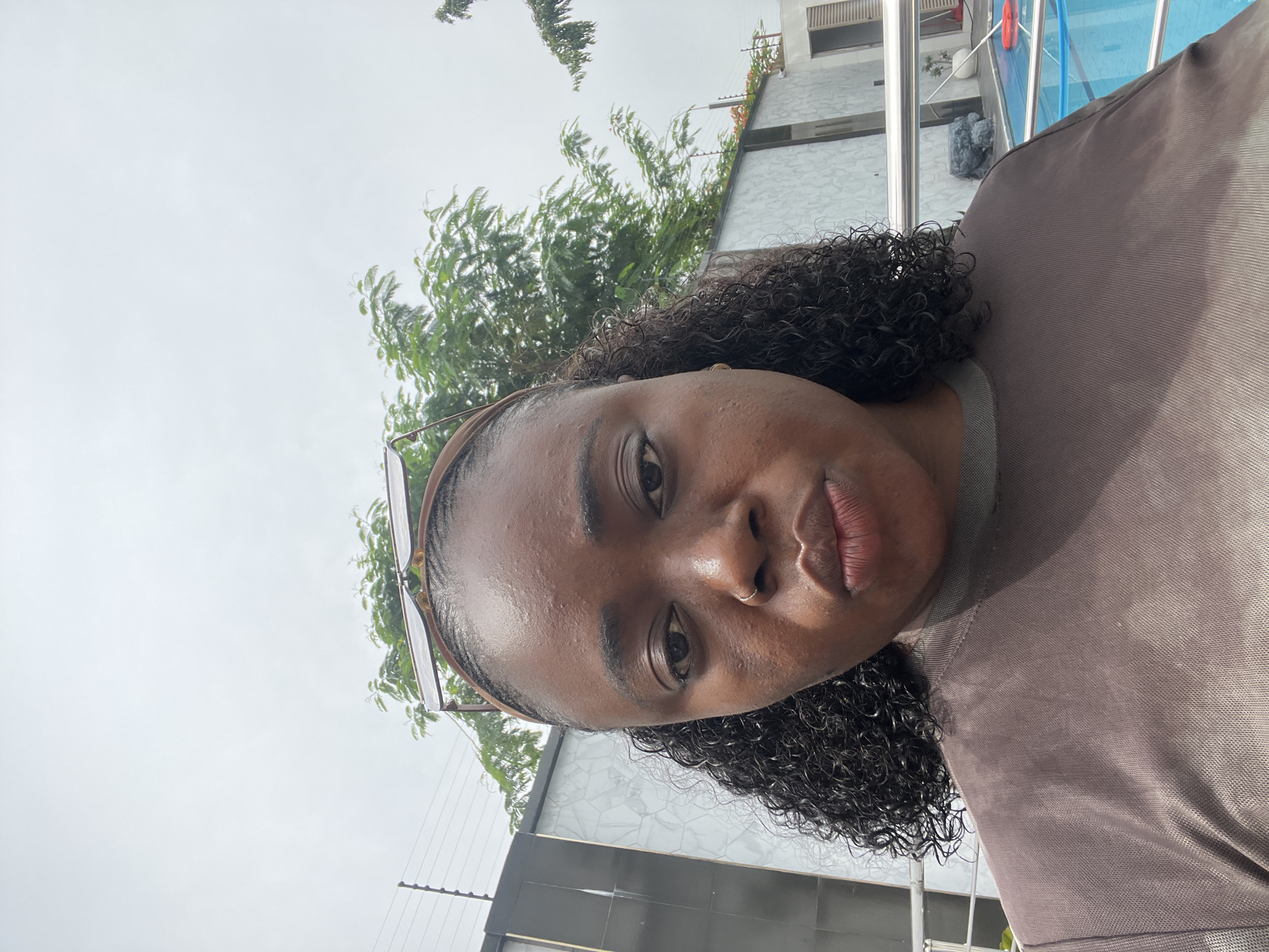 A medium shot shows a Black woman with curly hair pulled back by a headband, wearing sunglasses on top of her head. She is looking directly at the camera with a neutral expression. She wears a brown shirt. Her face is slightly angled to her left, and the image is a selfie. The background includes a light-colored building with a textured surface, green trees, a swimming pool with blue water, and a metal railing. There is a fence with electric wires visible to the left of the building. The sky is overcast. The location is identified as Abuja, Nigeria.