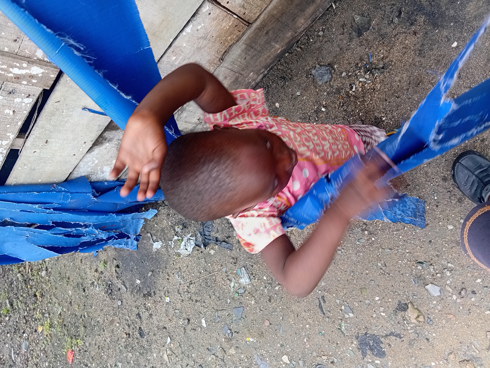 An overhead medium shot captures a young child in the city of Unknown, Unknown, engaged with a torn blue tarp. The child, wearing a pink and white patterned shirt, is seated or crouched on a ground composed of dirt, small stones, and scattered debris. The blue tarp, a significant element, is ripped and frayed, with the child actively pulling at it. To the right of the child, a wooden structure with weathered planks is visible. The lighting suggests an outdoor environment, possibly during daylight. The overall scene depicts a candid moment of a child interacting with their surroundings in what appears to be an urban or semi-urban setting.
