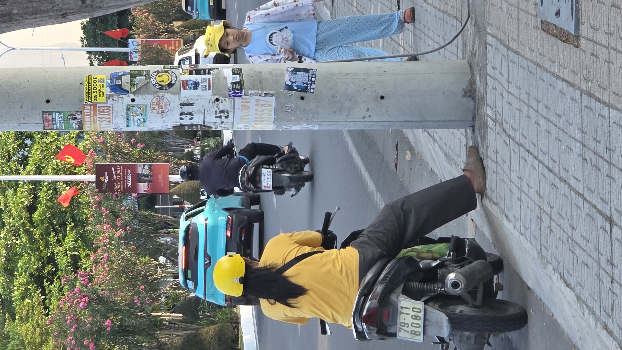 This image captures a vibrant street scene, likely in Bắc Nha Trang, Vietnam, characterized by common urban elements and local culture. The perspective is tilted, showcasing a sidewalk, a road, and the immediate surroundings.
In the foreground, a person with long dark hair, wearing a yellow shirt and a bright yellow helmet, is seated on a scooter or motorcycle, facing away from the viewer. Their license plate reads "79-T1 8080". Further down the road, another individual on a motorcycle, dressed in dark clothing and a black helmet, is visible with license plate "79-21 1569". A light blue car is also part of the traffic flow.
On the sidewalk, a young child wearing blue pajamas with a cartoon character and a yellow hat stands beside a large concrete utility pole, looking towards the observer. The pole itself is a focal point, heavily adorned with a variety of stickers and posters. These include advertisements in both Vietnamese and Russian, such as "МЕЖДУНАРОДНЫЕ ПРАВА ЗА 1 ДЕНЬ за 5000 р" (International driving permits in 1 day for 5000 p), "АРЕНДА БАЙКОВ" (bike rental), "ОБМЕН ВАЛЮТ" (currency exchange), "№1 ШАШЛЫК BBQ" (No. 1 BBQ), and "МАЧ-ТЕРАПЕВТ" (Match Therapist). Vietnamese text like "TIÊN, CẦM ĐỒ" (pawn shop) and "NHỮNG KỲ QUAN" (wonders) also appears, along with QR codes and other graphical elements.
The roadside is lush with green trees and bushes featuring pink flowers. Several red flags, some displaying the hammer and sickle emblem, are visible among the foliage, adding a distinct cultural identifier. The setting appears to be daytime, with soft, natural lighting. The blend of Vietnamese and Russian text on advertisements is a strong indicator of Nha Trang's status as a popular tourist destination for Russian visitors.