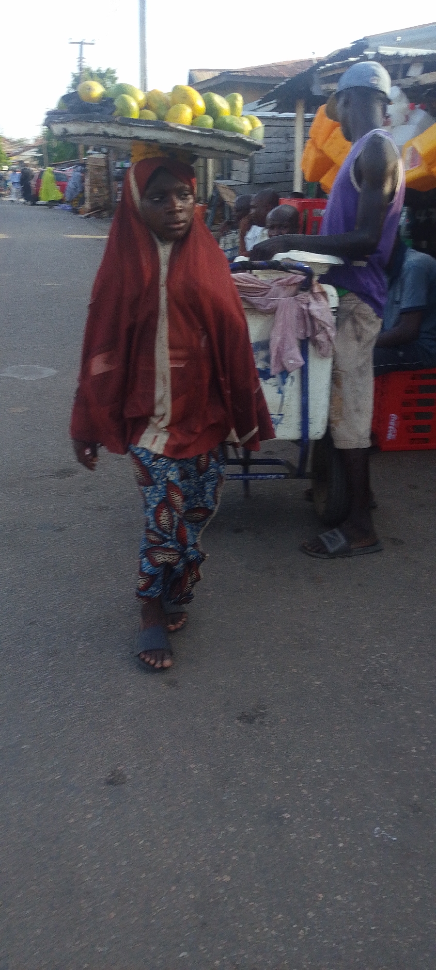 The image depicts a woman in Lokoja, Nigeria, carrying a tray of fruits on her head. She is wearing a red headscarf, a light-colored top, and patterned blue and red pants. She is wearing open sandals. The ground is asphalt, and the surrounding environment suggests a street scene with vendors. A man is standing beside a white cart with a blue frame. He wears a purple tank top, grey shorts, and a light blue hat. A red crate with "Coca-Cola" branding is visible behind the man. A second man is seated behind the cart. The scene is lit by natural daylight.