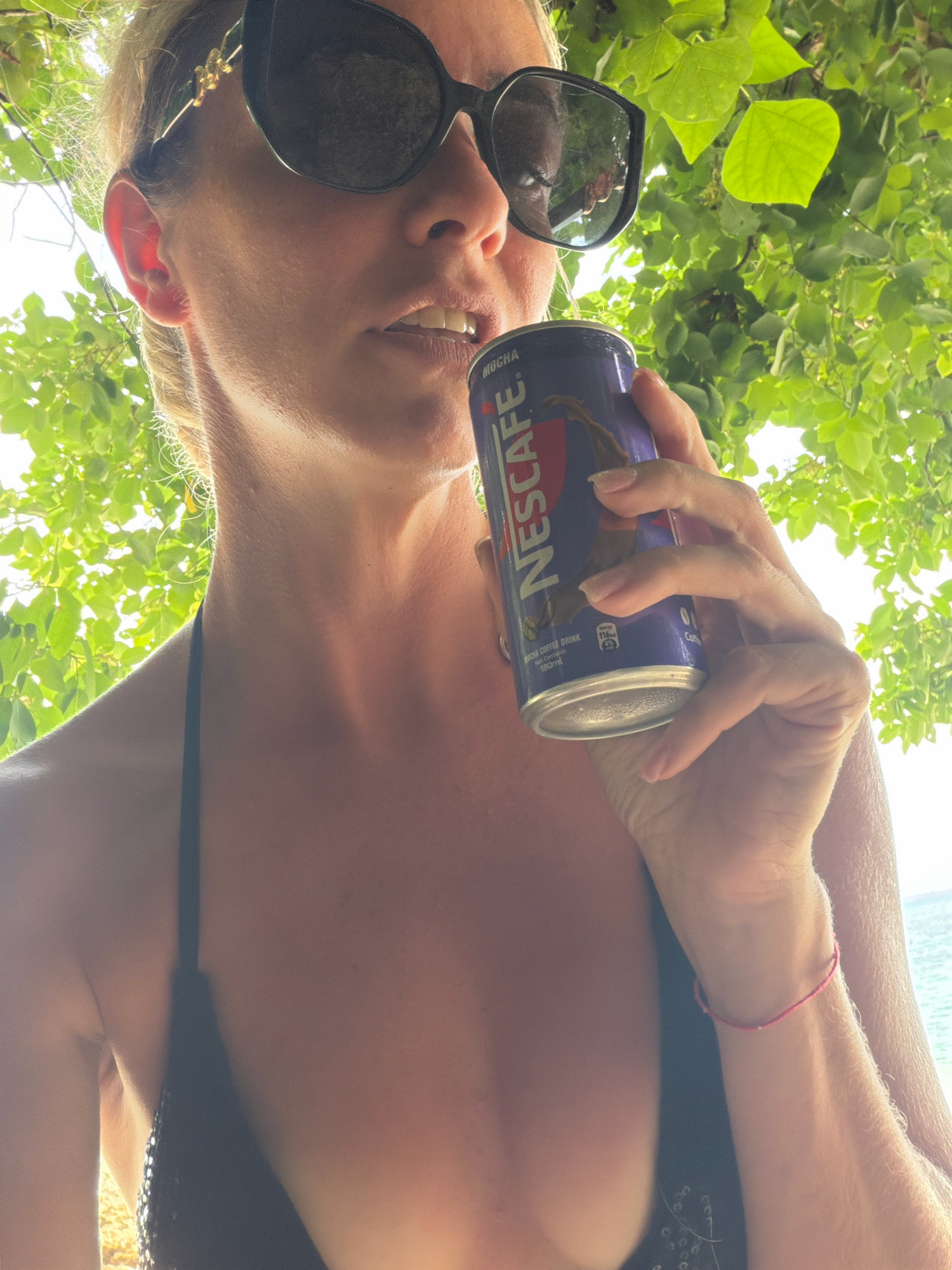 A medium shot captures a woman from the chest up, wearing a black bikini top and large sunglasses, holding a can of Nescafé Mocha coffee drink to her lips. She is positioned outdoors, with lush green foliage framing the top of the image, suggesting a natural, possibly tropical environment. The background shows a hazy glimpse of what appears to be the ocean, indicating a coastal setting. The lighting is bright and natural, with sunlight filtering through the leaves, creating a dappled effect. The woman's skin appears tanned, and her hair is blonde and pulled back. The overall scene evokes a relaxed, outdoor, and possibly vacation-like atmosphere. The location is identified as Malay, Philippines.