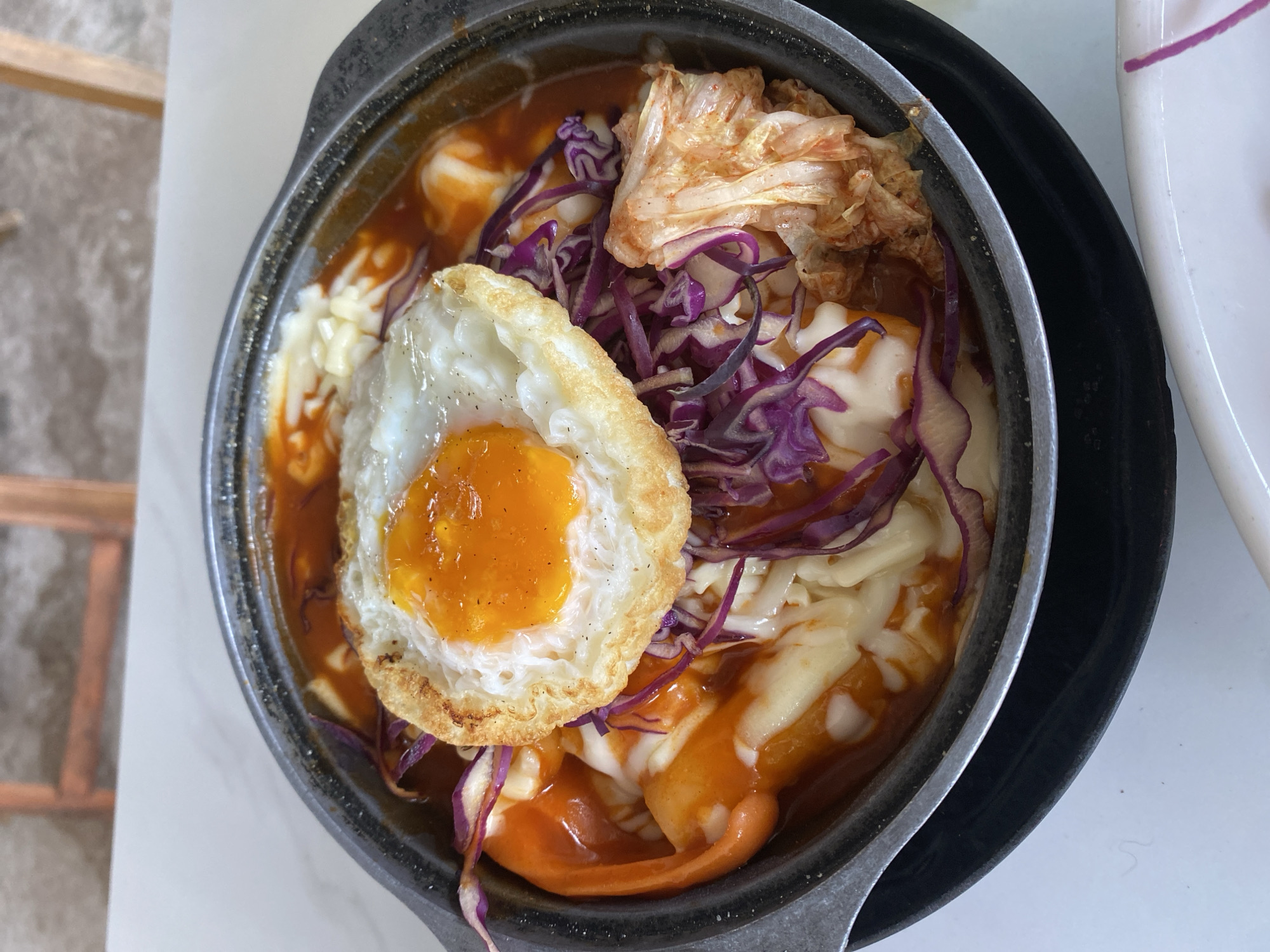 The image shows a bowl of Tteokbokki with a fried egg, shredded mozzarella cheese, purple cabbage, and kimchi. It is served in a metal pot on top of a black plate. The picture was taken in Mũi Né, Vietnam. There is a wooden chair leg visible at the edge of the frame. The table is white marble.
