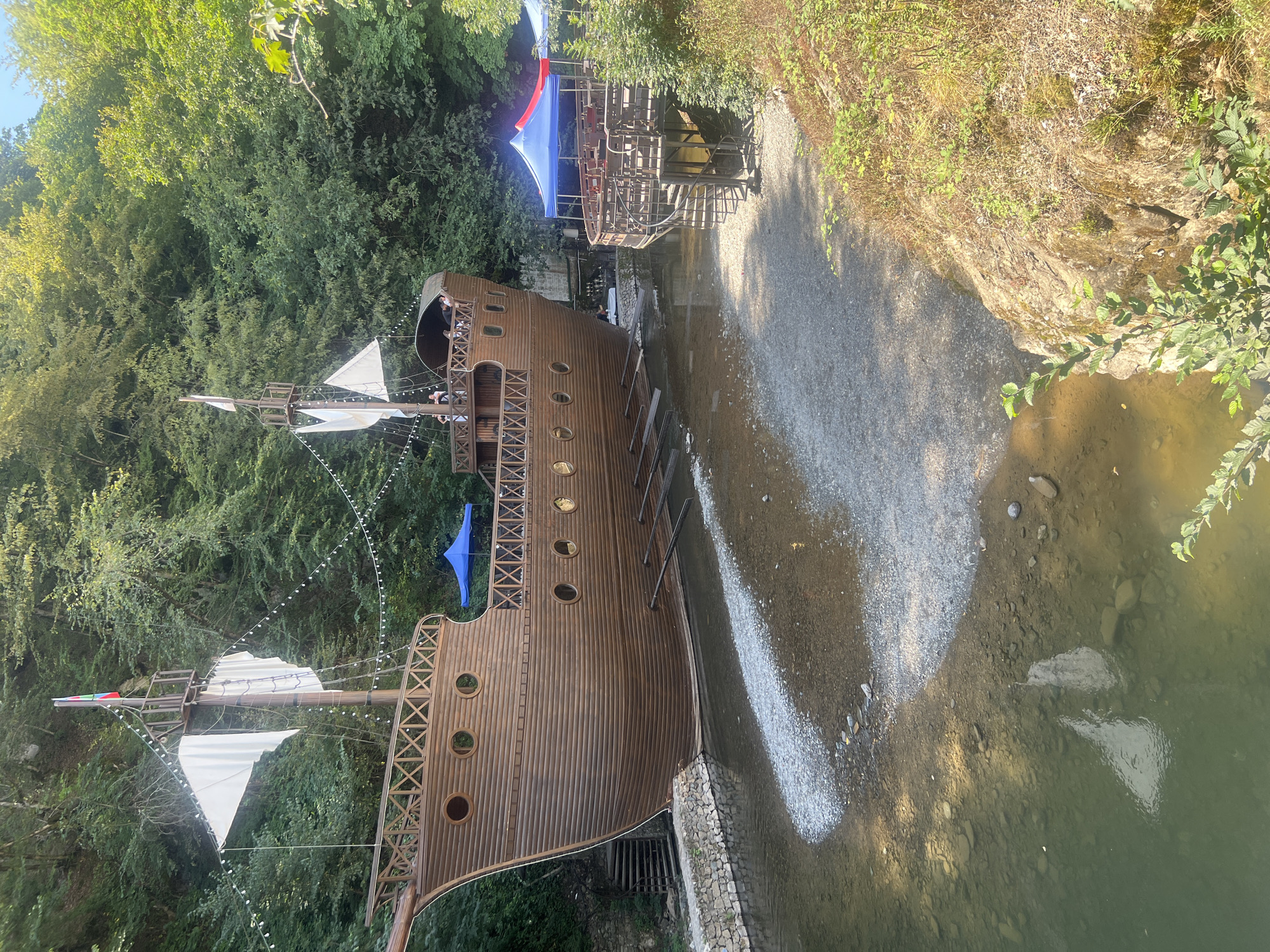 The image depicts a large, wooden, pirate ship replica situated on the banks of a clear, shallow river in Sarısa, Azerbaijan. The ship is the primary subject, appearing to be a restaurant or entertainment venue, with tables and seating visible on its deck and on a nearby elevated platform. Two tents, one blue and another red and blue, are set up near the platform, providing shade for outdoor seating. The ship has multiple levels, a mast with sails, and decorative windows. A spiral staircase connects the riverbank to the elevated platform. The riverbed is visible, covered with stones and pebbles, and the water reflects the ship and surrounding trees. Dense green foliage and trees line the riverbanks, suggesting a natural, forested environment. The sky is clear and bright.