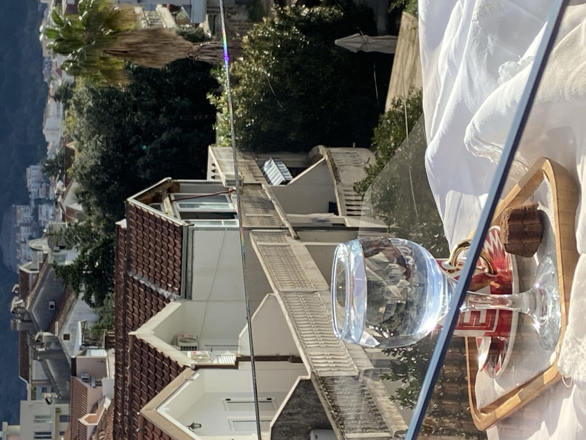 A close-up shot depicts a glass of water and a small, wooden tray with food atop a clear surface, likely a balcony, in Budva, Montenegro. The background reveals a dense cityscape with residential buildings featuring red-tiled roofs, interspersed with greenery and glimpses of a distant mountain. The structures exhibit varied architectural styles with balconies and windows visible. A faint vertical rainbow-like refraction occurs in the center background.