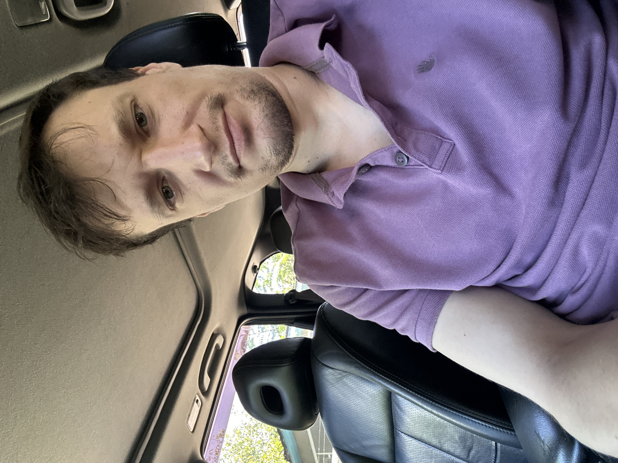 A man with light skin, brown hair, and a light beard is taking a selfie in the driver's seat of a car. He is wearing a purple polo shirt with a small logo on the left chest. The car's interior is visible, including the black leather driver's seat, a sunroof, and the exterior visible through the windows, which appears to be a bright, sunny day with some greenery. The man's face is slightly turned towards the camera, with a neutral expression.