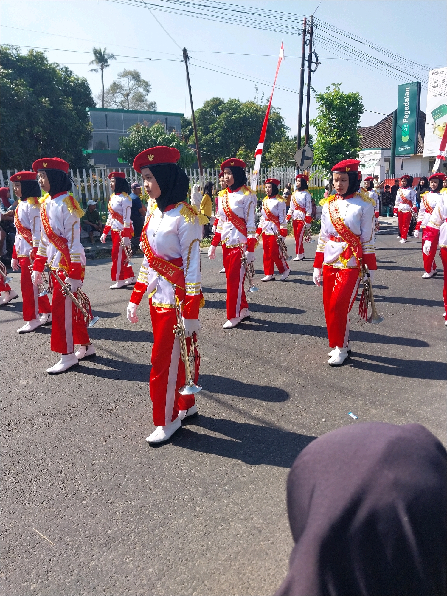 In the city of Nglegok, Indonesia, a marching band comprised of young women in uniform is participating in a parade. The band members, all wearing red berets, black headscarves, white shirts with gold trim, and red pants with white stripes, are holding brass instruments, specifically trumpets. They are marching on a paved road, with a line of spectators visible in the background. The environment includes trees, buildings, and a clear blue sky, suggesting a sunny day. A prominent green sign with the word "Pegadaian" is visible in the background, indicating a financial institution. The image captures a moment of community activity and celebration.