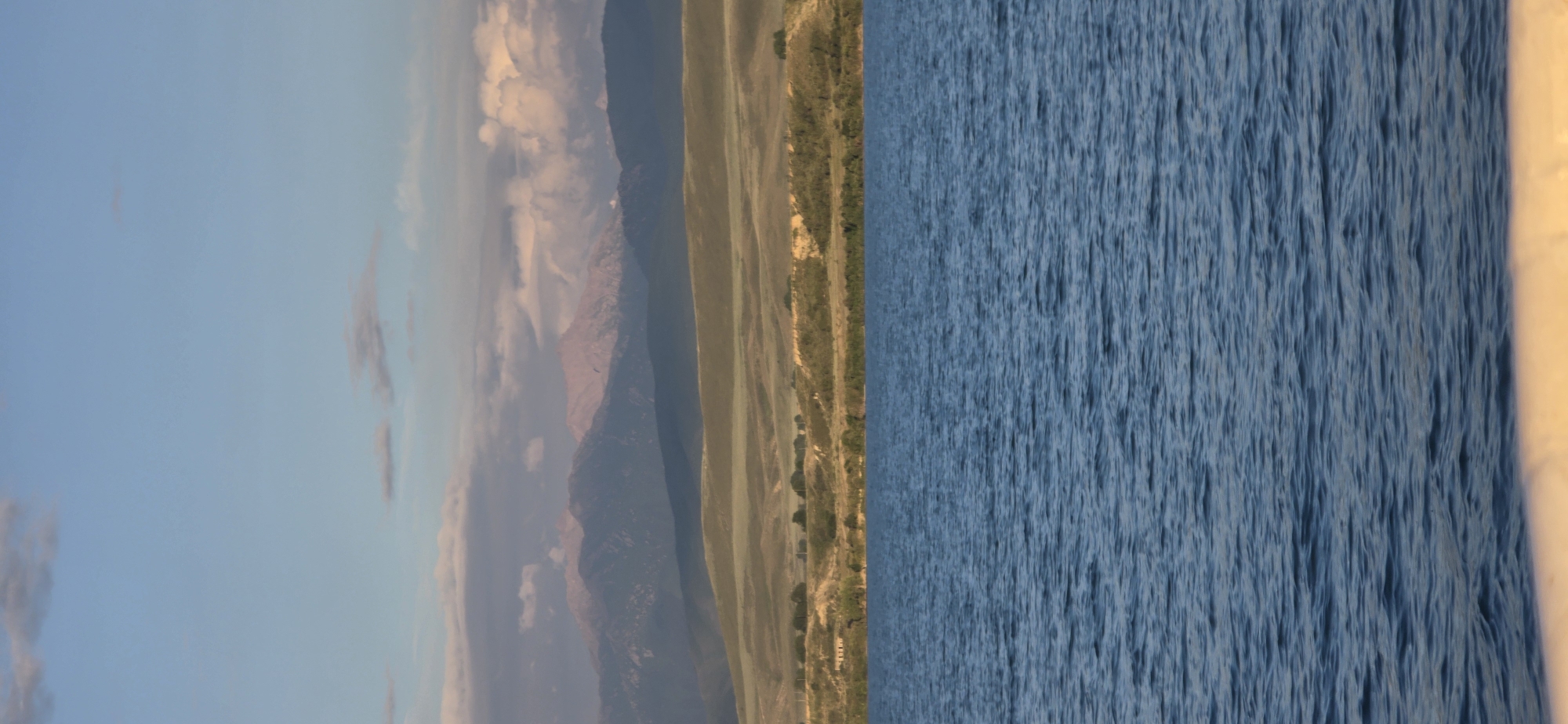 The image depicts a wide view of a body of water in the foreground, with a textured surface indicative of ripples. Beyond the water, a gently sloping, treeless landscape stretches towards a range of mountains in the distance. The mountains exhibit varied geological formations and are partially obscured by clouds. The sky above is a pale blue, with scattered, soft clouds that are illuminated by a warm light, suggesting either sunrise or sunset. There are no people or discernible man-made structures in the immediate vicinity of the water, nor in the middle ground landscape. The scene evokes a natural, serene environment. The location is not identifiable as Pristan'-Przheval'sk, Kyrgyzstan, based on the visual evidence presented in the image.