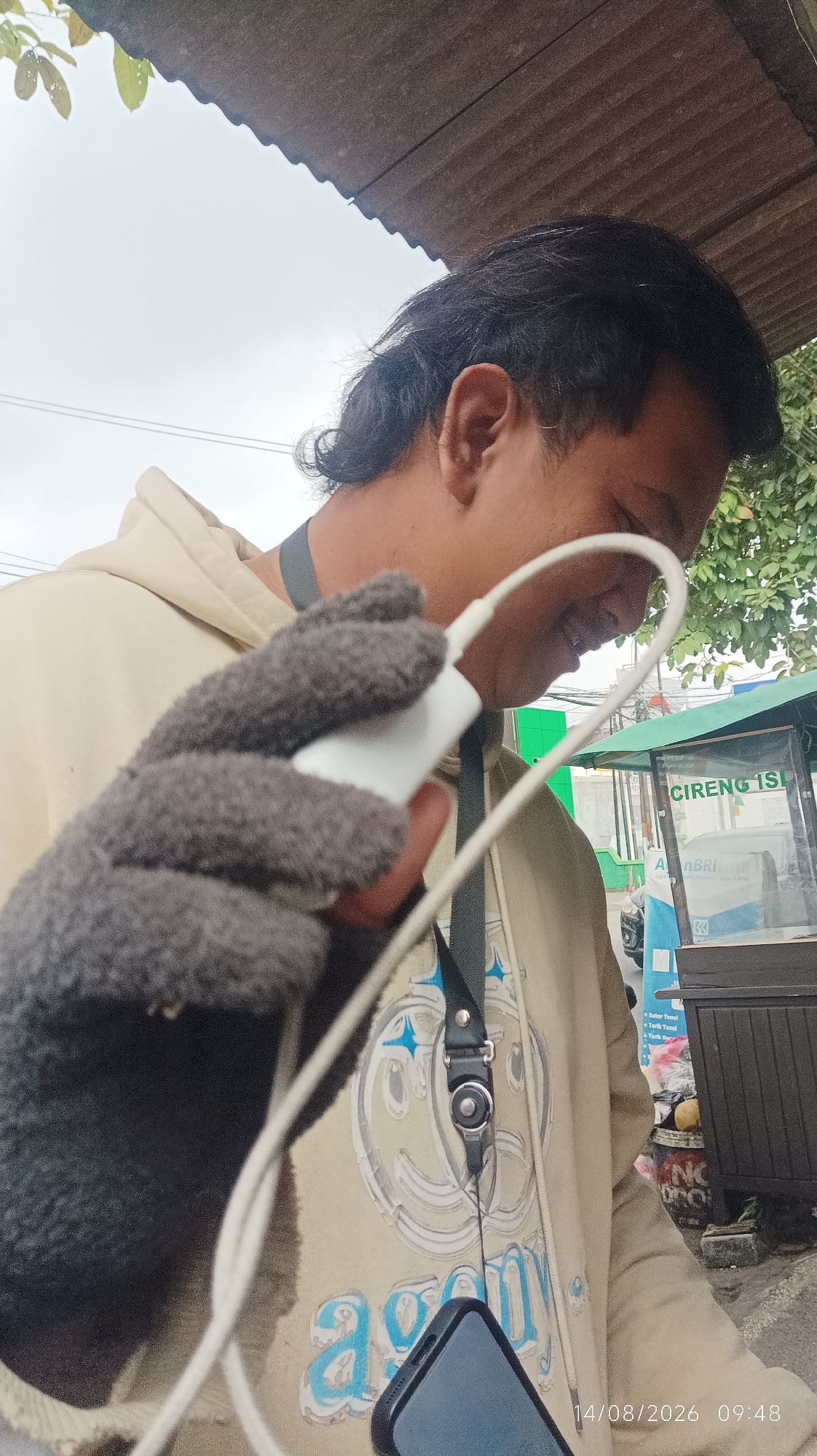 A person wearing a light brown hoodie and gray fuzzy gloves is holding a white charging brick connected to a white cable. The person's face is partially visible from the side, and they appear to be looking down at the charging brick. The background shows a cloudy sky, a corrugated roof, trees, and a food stall with a sign that reads "CIRENG ISI". The timestamp "14/08/2026 09:48" is visible in the lower right corner. The setting is likely an outdoor area in Tasikmalaya, Indonesia, given the presence of the food stall and the Indonesian text on the sign.