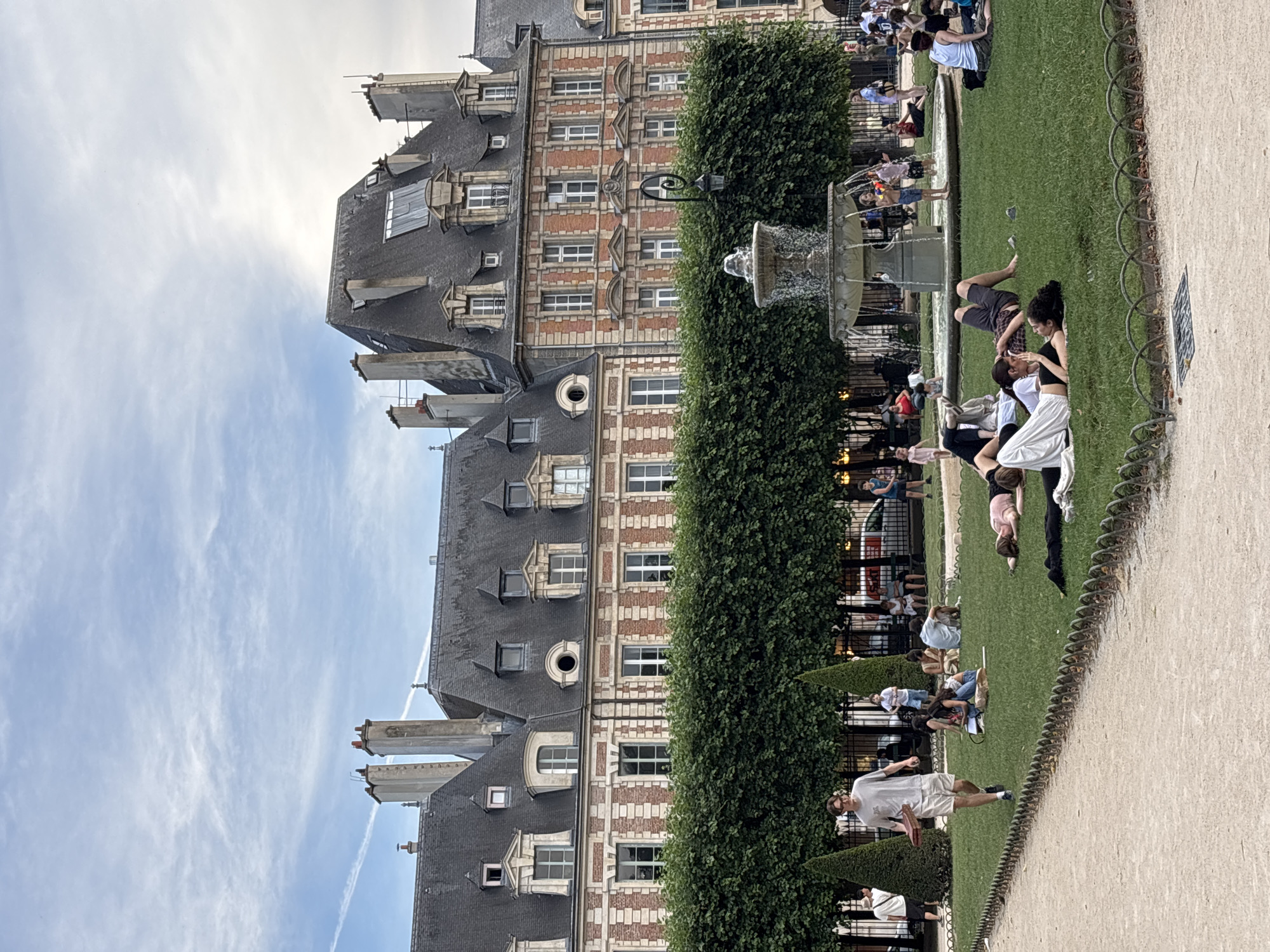 The image depicts the Place des Vosges in Paris, France, on a cloudy day. A man in a light-colored t-shirt and shorts walks across a grassy area, carrying a flat, rectangular object. Several groups of people are scattered across the lawn, some sitting and others lying down. In the background, a large fountain is active, with several children playing around it. Behind the fountain and the people, a manicured hedge forms a distinct boundary, beyond which a grand, historic building with a red brick facade and steeply pitched mansard roofs stands. The architecture of the building, characterized by its dormer windows and chimneys, is typical of Parisian urban design. The sky above is partially overcast, with wisps of clouds visible. The overall scene conveys a sense of leisure and public enjoyment in an urban park setting.