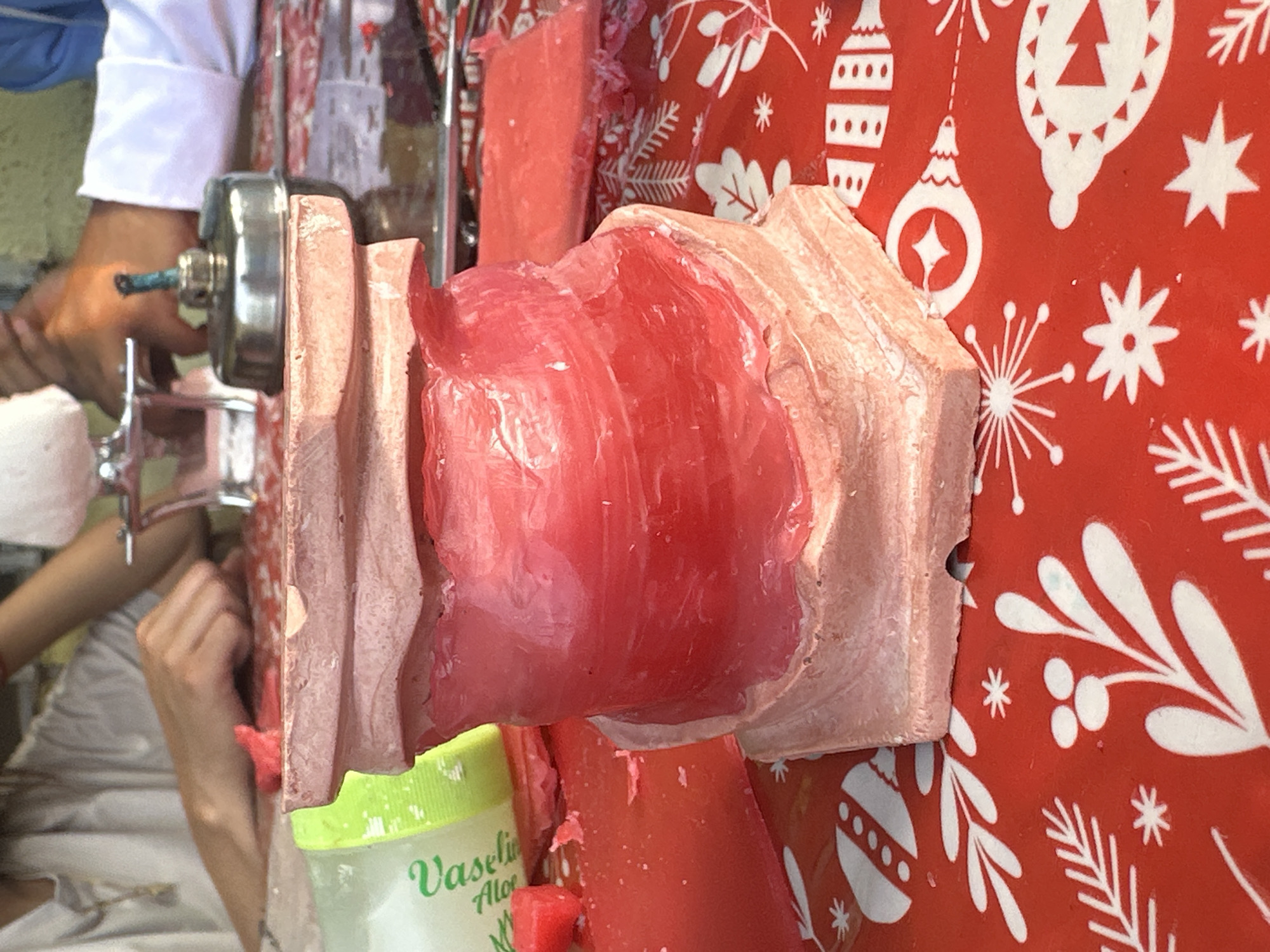 The image shows a process of making wax candles. A block of pink wax is being compressed through a shaping device, likely a manual press, which has a silver handle. There is a person in the background operating the press, wearing a white sleeve. A jar of Vaseline Aloe Fresh is sitting near the wax block. The entire scene is set on a red surface printed with white Christmas-themed designs such as ornaments, snowflakes, and trees. The setting appears to be indoors, possibly a workshop or studio. This is in Parroquia Ocumare, Venezuela.