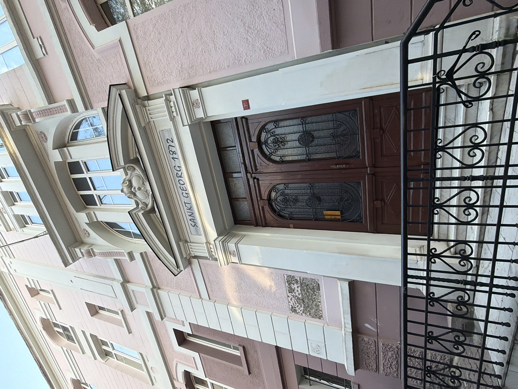 The image shows the entrance to a building with a pink facade. The entrance features a large brown double door with decorative metal grilles on the glass panels. Above the door, a stone inscription reads "SANKT GEORG 1872". The entrance is flanked by white pillars. A black metal railing with ornate scrollwork is in the foreground, leading up to a set of marble steps. The building has multiple windows with white frames. There is a small red sign with white text, reading "7b", on the right pillar. The sky is visible at the top of the image. The overall scene depicts the architectural facade of a historical building.
