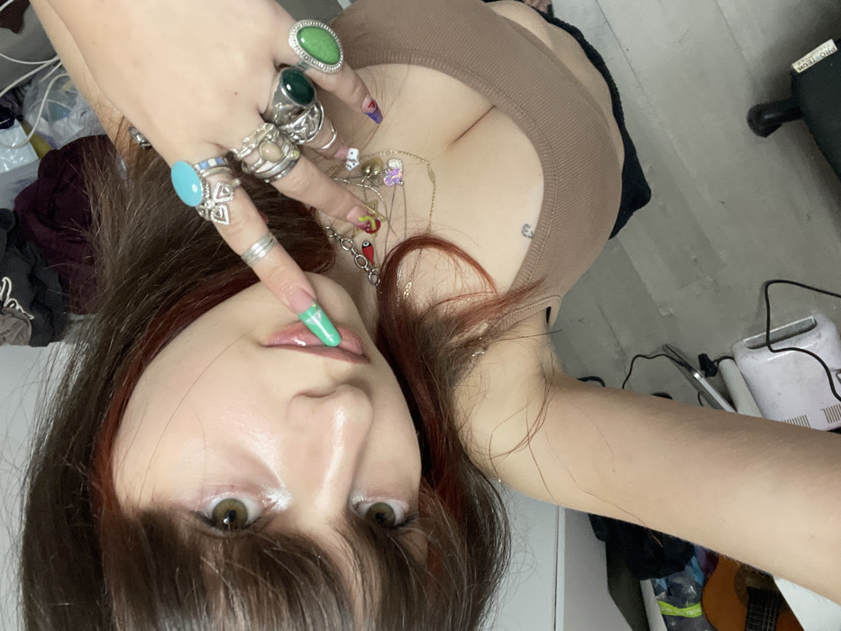 A woman with dark hair and light eyes poses in a selfie. She wears a brown top and multiple rings, including ones with large gemstones. Her nails are painted with colorful designs and one is green and touching her glossy lips. She also has multiple necklaces. In the background is a messy room with a guitar, a fan, and clothing.