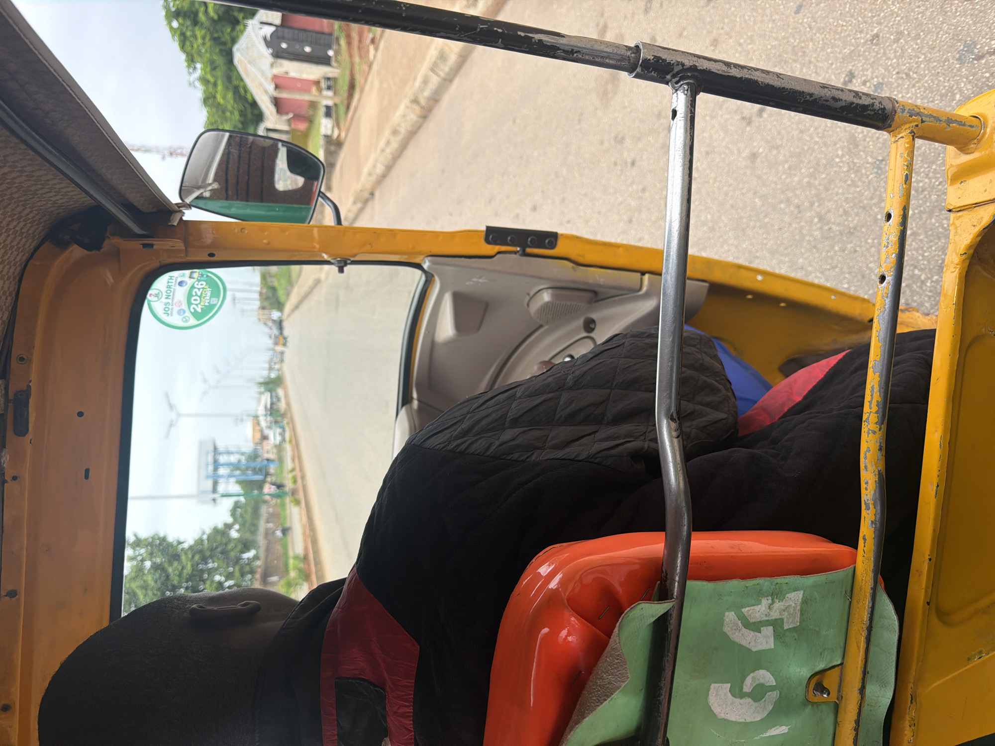 The image shows the interior of a yellow auto-rickshaw, also known as an "okada" or "keke napep," in Bukuru, Nigeria. The driver, a man with dark skin and short hair, is visible from the back, wearing a black quilted jacket with red accents. In the foreground, a green seat with the number "64" stenciled in white is visible, with an orange-colored object resting on it. The auto-rickshaw appears to be in motion, traveling down a paved road lined with trees and buildings. A large water tower is visible in the distance, along with streetlights. A circular sticker with the text "JOS NORTH 2026 TRICYCLE PERMIT" is affixed to the windshield. The sky is overcast.