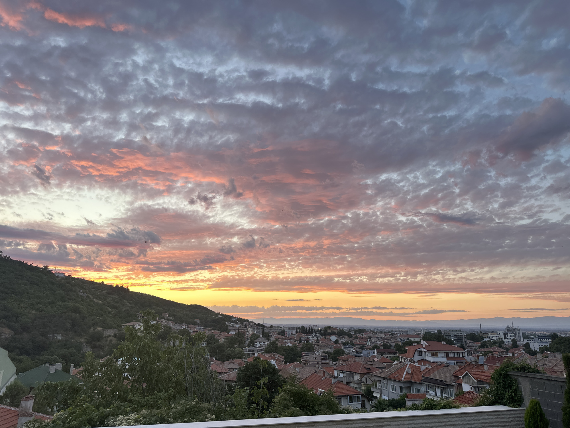The image depicts a panoramic view of the city of Asenovgrad, Bulgaria, at sunset. The sky is filled with dramatic clouds, painted in hues of orange, pink, and purple, with hints of yellow near the horizon. The setting sun casts a warm glow over the landscape.
In the foreground, the roofs of houses with red tiles are visible, interspersed with green trees and foliage. To the left, a densely wooded hillside slopes down towards the city. The middle ground shows a cluster of residential buildings, characteristic of a European town. In the distance, the city extends towards the horizon, with a mix of low-rise and some taller modern buildings. The sky transitions from a bright, sunlit band near the horizon to darker, more muted tones overhead. Mountain ranges are faintly visible in the far distance, silhouetted against the twilight sky. The overall scene conveys a sense of tranquility and the beauty of a natural phenomenon over an urban setting.