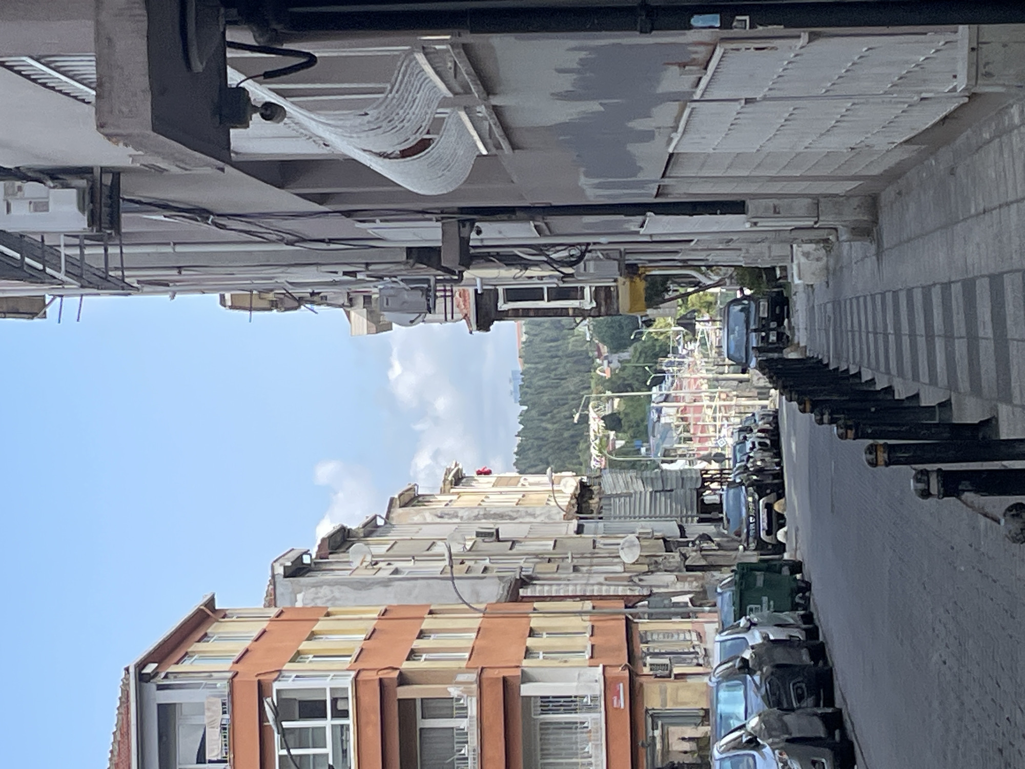 The image depicts a narrow, uphill street in Kadıköy, Turkey, lined with apartment buildings. The buildings are a mix of styles and colors, with the one on the left being orange and yellow, and the one on the right being a neutral gray. Several cars are parked along the street, and a green dumpster is visible. The sidewalk on the right has a distinctive black and white striped pattern. In the distance, a dense forest covers rolling hills. The sky is blue with a few scattered clouds. The overall impression is of a typical urban street scene in a Mediterranean climate.