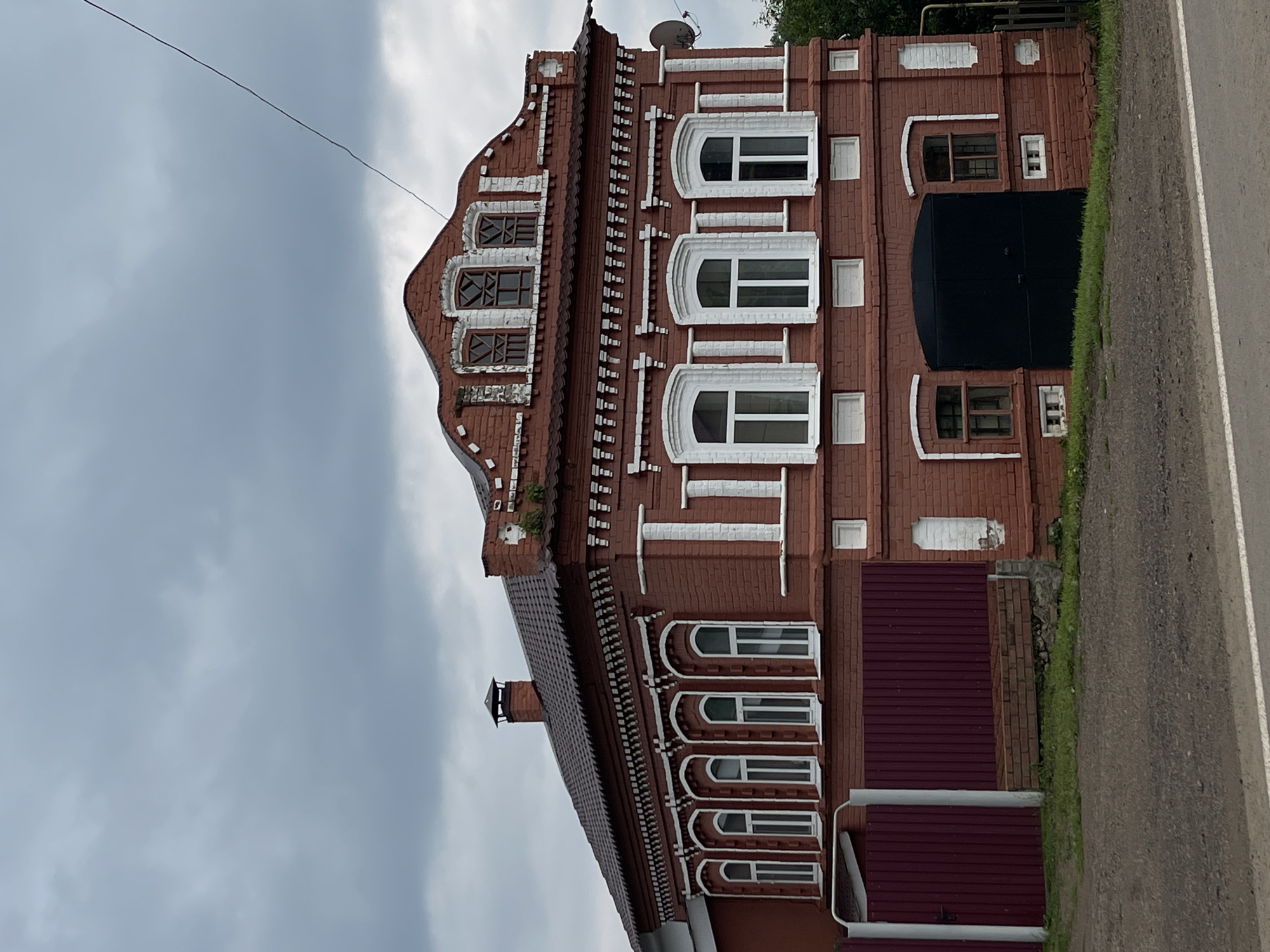 A two-story red brick building with white trim and dark brown roof is viewed from a slightly low-angle, side-on perspective. The building has a symmetrical facade with a central gable. The first floor has a large black garage door flanked by two windows, each with a white arched trim. The second floor features three large arched windows with white trim, and above them, within the gable, are three smaller, triangular-shaped windows. The building is bordered by a dark red corrugated metal fence on the left side. The sky is overcast with gray clouds. The ground in front of the building is covered with patchy grass and gravel. A paved road runs across the bottom of the image.