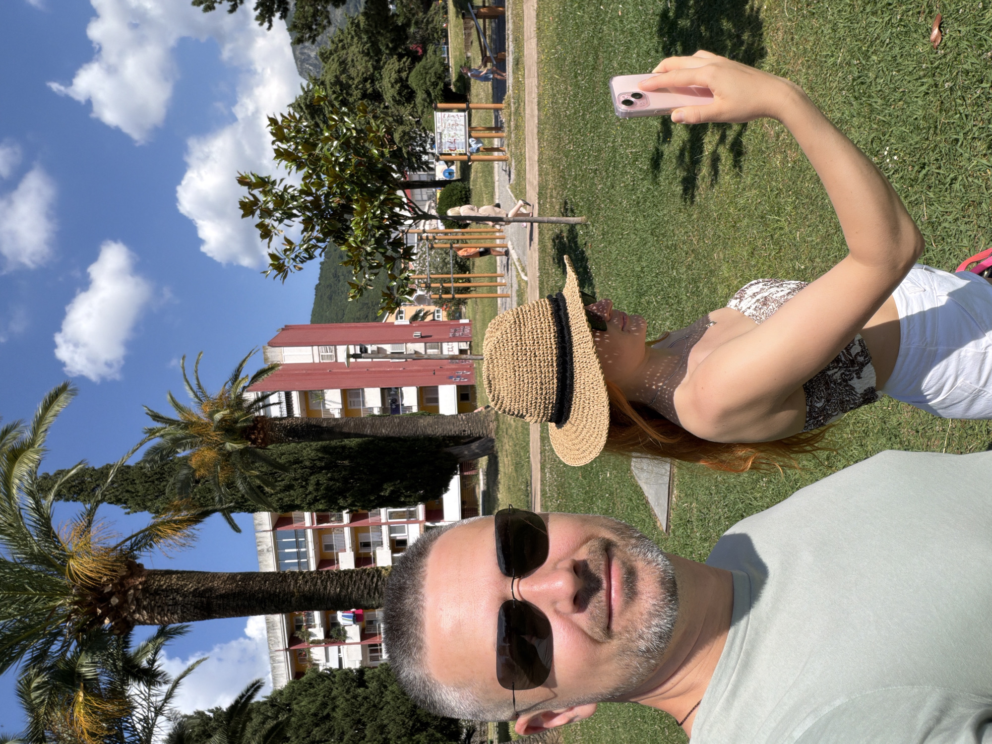 The image displays a sunny outdoor selfie featuring two individuals in a park-like setting, consistent with Budva, Montenegro. In the foreground, a man with short grey hair and a beard, wearing dark sunglasses and a light green t-shirt, smiles faintly at the camera. Slightly behind and to his right, a woman with reddish-brown hair, wearing a straw hat with a black band, sunglasses, and a patterned top, is holding a light pink smartphone to capture the photo. White shorts are visible on the woman.
Behind the couple, a green grassy area is visible, dotted with large palm trees and other dense green trees. In the mid-ground, there's a children's playground with wooden structures, where several indistinct figures can be seen. Further back, multi-story residential buildings with red roofs and balconies stand among the trees. Majestic mountains rise in the far background under a bright blue sky adorned with fluffy white clouds. The clear skies and strong sunlight indicate it is daytime, likely a pleasant morning or afternoon. The overall scene conveys a relaxed and enjoyable atmosphere. No discernible text is visible on any signs or objects.