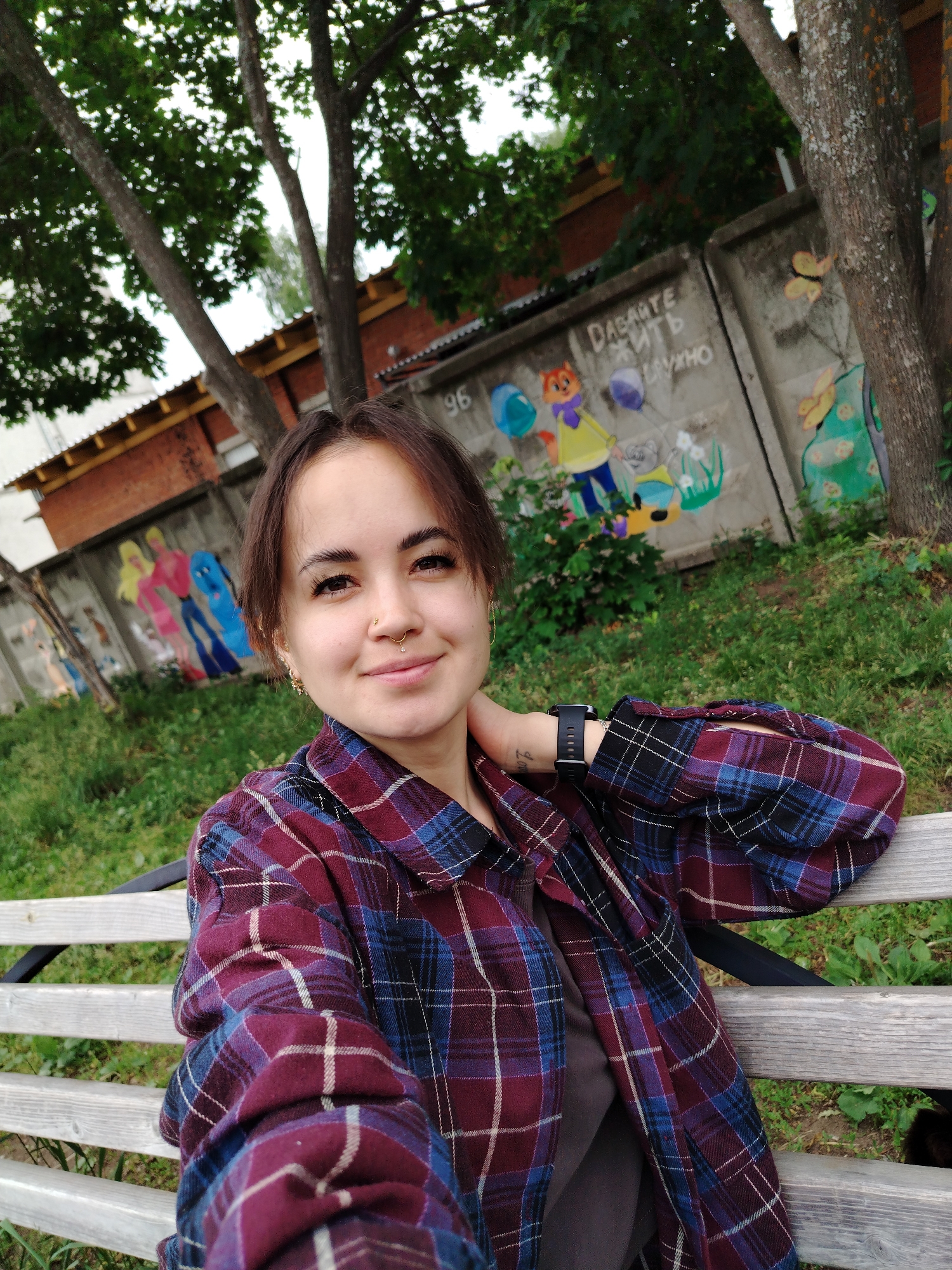 A young woman with dark hair, styled in a bob with bangs, is seated on a wooden bench outdoors. She is wearing a maroon and blue plaid flannel shirt over a dark gray t-shirt. She has a nose ring with a small jewel and an earring in her left ear. Her right arm is resting on the back of the bench, and her left hand is touching her neck. A black smartwatch is visible on her left wrist. In the background, there is a concrete wall with colorful graffiti depicting cartoon characters and butterflies. Behind the wall, a brick building and trees are visible. The ground is covered with green grass and some scattered plants. The scene appears to be a public outdoor space, possibly a park or a courtyard, located in Noviy, Russia.