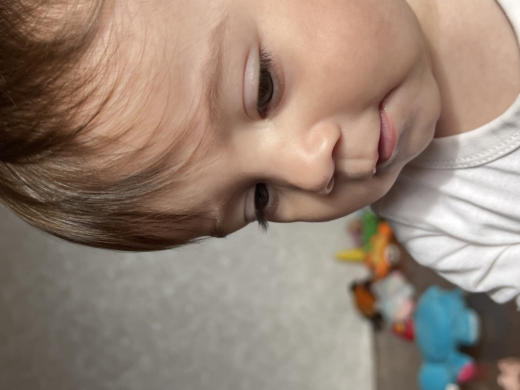 A close-up, eye-level portrait captures an infant in what is stated to be a location in Elena, Bulgaria. The child, with fair skin, light brown hair parted to the left and swept across the forehead, and dark brown eyes, is positioned slightly right of center, gazing downwards and to the right. The mouth is slightly open, with a visible trace of moisture on the lower lip. The infant wears a white, short-sleeved garment. The background is heavily blurred: a light gray, textured wall is visible in the upper left, while a darker surface, possibly a floor, occupies the lower background. Scattered on this surface are multiple indistinct, brightly colored children's toys, including a prominent blue, rounded object on the far left, and other orange, yellow, and pink forms further back. The lighting is soft and even, illuminating the child's face.