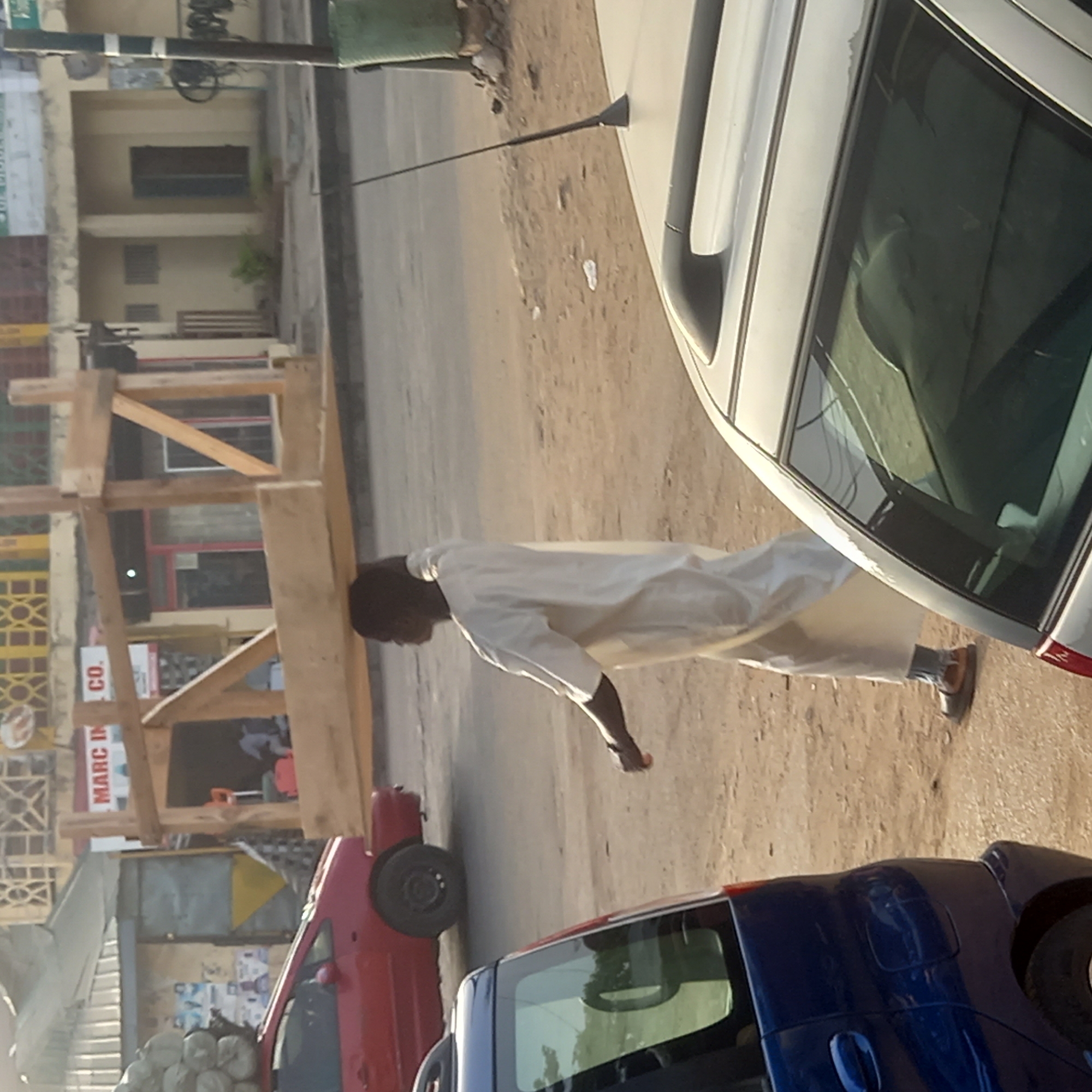 The image presents a daytime street scene in Dadi Riba, Nigeria, viewed from a slightly elevated perspective, possibly from inside a vehicle.
In the center of the frame, a man is walking or standing on the unpaved shoulder of the road. He is wearing a light-colored, long-sleeved tunic (possibly white or off-white) and matching trousers, with dark sandals on his feet. His head is turned slightly to his right.
The street features a paved section in the middle, flanked by wider, unpaved dirt or sand shoulders. Several cars are visible. In the right foreground, a light-colored sedan (possibly silver or white) is prominently featured, showing its side and rear. Another vehicle, a blue car, is in the bottom right corner. A red car is partially visible in the lower left mid-ground.
Along the left side of the street, there are buildings. A substantial wooden scaffolding structure, possibly for construction or repair, stands in front of one of these buildings. On the building behind the scaffolding, a sign with the text "MARC IN CO." is discernible. Further along the street, more building facades with windows and structural details can be seen. In the lower-left portion of the image, several white sacks, possibly containing goods or building materials, are stacked near the roadside.
The lighting suggests a sunny day, with warm hues that could indicate either morning or late afternoon. The overall impression is that of a typical urban or semi-urban street, busy with daily activities.