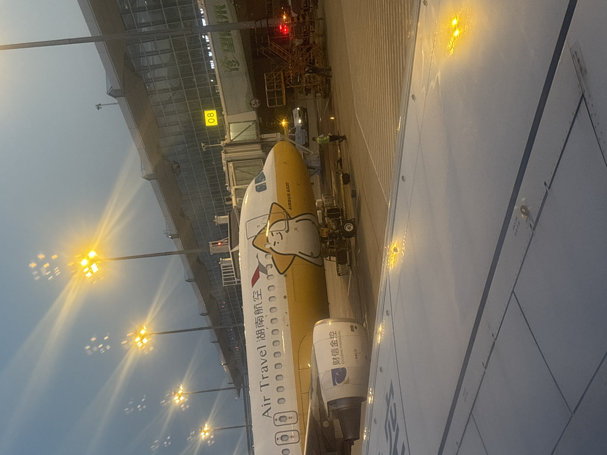 The image depicts an airplane at an airport gate. The airplane's livery is predominantly white and yellow, with a cartoon character adorning the tail. The text "Air Travel" and "湖南航空" are visible on the fuselage. The airplane is connected to the terminal via a jet bridge, labeled with the number "08". Several individuals are present on the tarmac, including airport personnel in high-visibility vests. Various airport vehicles and equipment, such as a baggage cart and stairs, are also visible. The airport terminal is a modern structure with a glass facade. The scene is illuminated by runway lights and the ambient light of dusk. The image is taken from the perspective of someone inside another airplane, looking out at the scene.
The provided context indicates the location is Xinwu District, China. Xinwu District is a district of Wuxi city in Jiangsu province. The airport in Xinwu District is Sunan Shuofang International Airport (WUX).