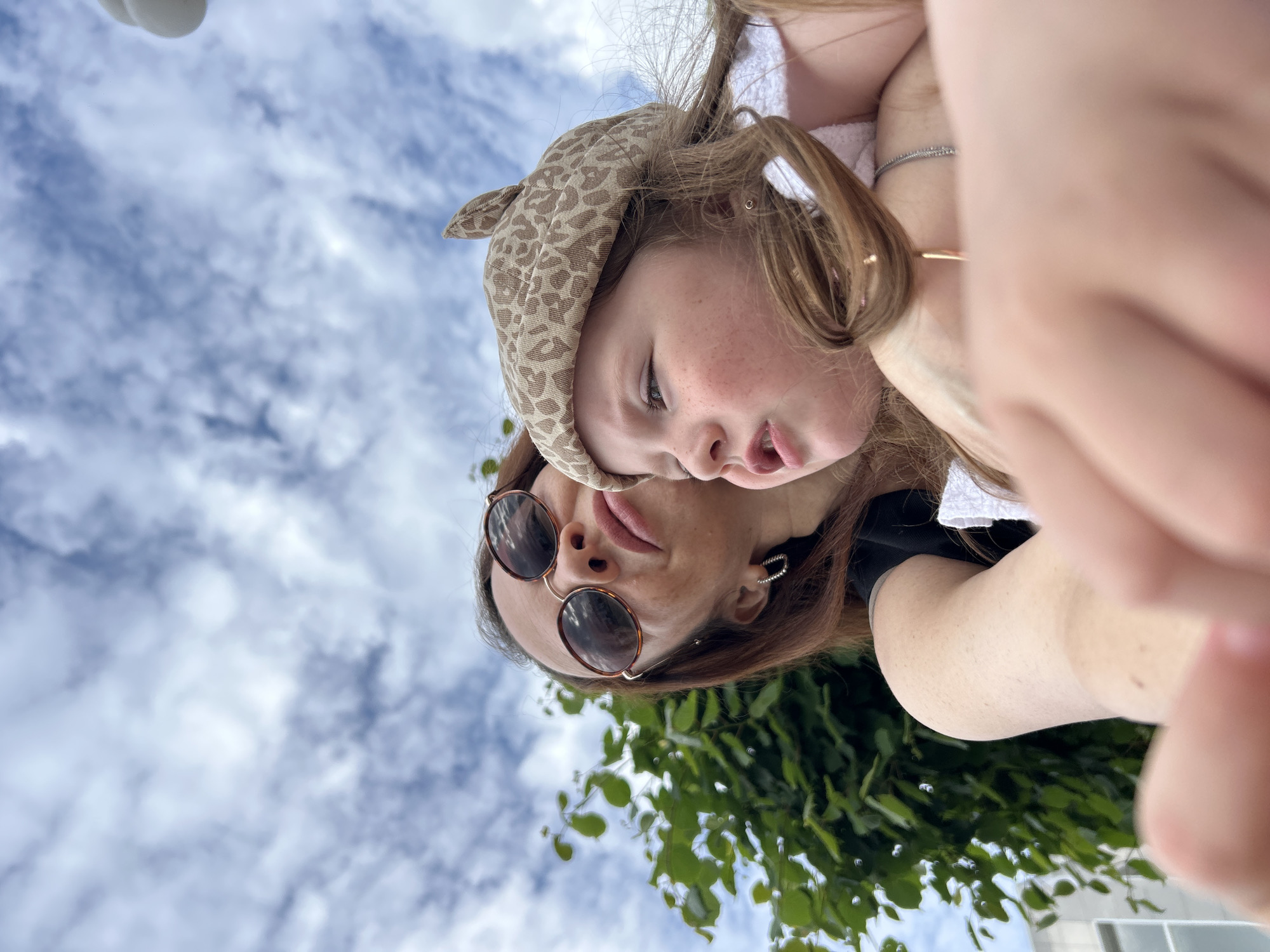 A low-angle, close-up selfie shows a woman and a young girl outdoors on a cloudy day. The woman, wearing sunglasses and hoop earrings, has reddish-brown hair and is looking upwards. The girl, wearing a tan beret with a leopard print pattern, is to the woman's right and looking to the side with her mouth slightly open. Green foliage is visible to the left of the frame, and a building can be seen in the background. The setting is consistent with an outdoor urban environment, such as a park or street in Chaikovsky, Russia.