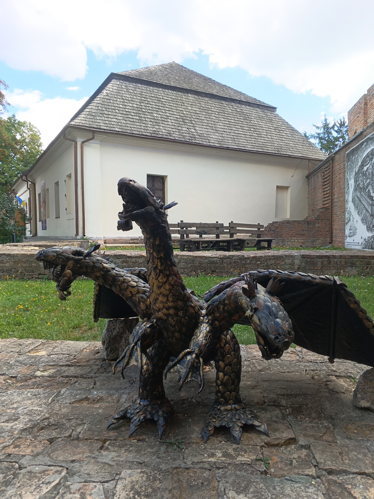 The image shows a three-headed dragon sculpture made of metal, positioned on a stone surface in front of a building. The sculpture is detailed, with scales and claws clearly visible on the dragons. In the background, there is a light-colored building with a grey, shingled roof. To the right of the building, a brick wall is visible, with a large black and white mural or poster attached to it. There are trees and green grass with yellow wildflowers in the foreground and midground. The sky is blue with some white clouds. This sculpture is located in Lubart's Castle in Lutsk, Ukraine.