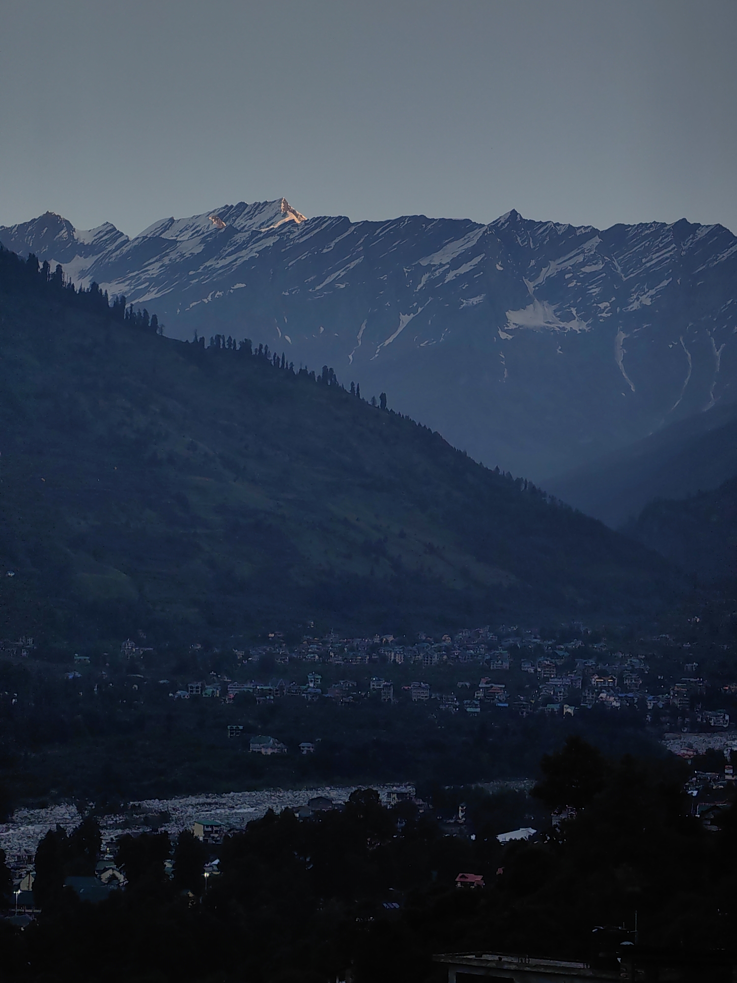The image depicts a panoramic view of the mountainous region near Vashisht, India, at dusk. The sky is a gradient of pale blue to dark blue, with the last rays of sunlight illuminating the snow-capped peaks in the distance. A prominent mountain peak in the center of the upper frame catches a golden hue, suggesting the setting sun. The mid-ground is dominated by lush green, heavily forested hillsides that slope down towards a valley. A small town, identified as Vashisht, is nestled within this valley, with clusters of buildings and houses visible. The town appears to be situated near a river or stream, indicated by a rocky, wide channel in the foreground. The foreground is heavily shadowed, with dark silhouettes of trees and structures obscuring the details of the immediate surroundings. The overall scene is serene and captures the transition from daylight to night in a mountainous landscape. There are no visible people or specific actions taking place.