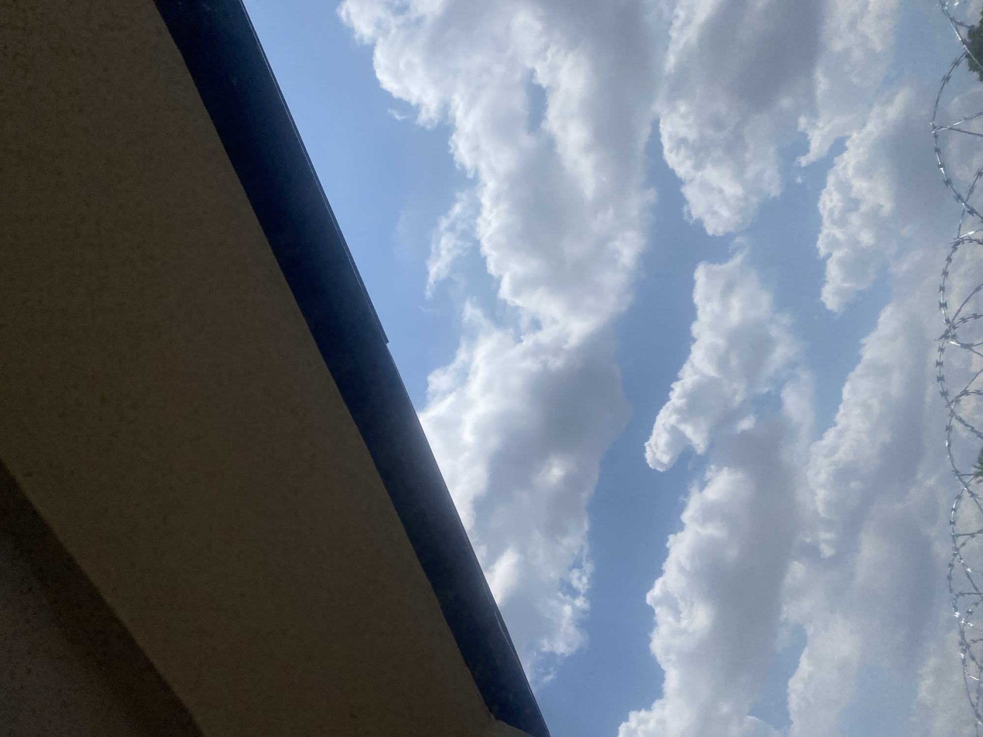 The image displays a view of the sky from underneath an overhang, likely a roof or balcony. The sky is a bright blue with scattered white cumulus clouds. In the foreground, at the bottom of the image, a coiled barbed wire fence is visible. The scene appears to be outdoors. There are no people or animals depicted in the image. The environment suggests an urban or semi-urban setting, possibly a perimeter fence in the city of Lafia, Nigeria, given the context.