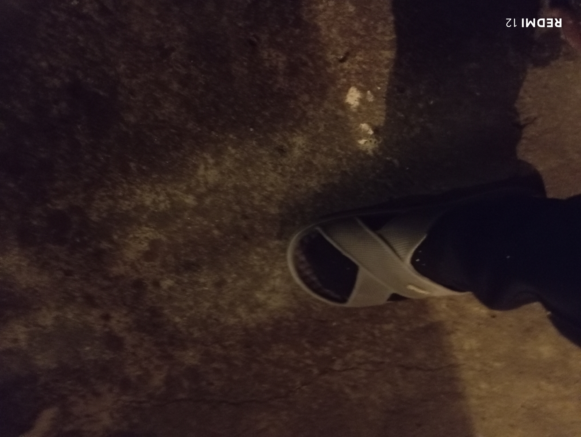 The image is a close-up, low-angle shot of a person's foot wearing a gray sandal with two crisscrossed straps. The foot is clad in dark-colored clothing, possibly a sock or the lower part of a pant leg. The sandal is positioned on a textured, dark gray surface, which appears to be asphalt or concrete with some lighter colored debris scattered on it. The lighting is dim, casting shadows and highlighting the texture of the ground. The "REDMI 12" watermark is visible in the bottom left corner. The scene is outdoors, potentially in a city area like Aygedzor, Armenia, given the context provided. The overall impression is gritty and urban, with a focus on the footwear and the ground.