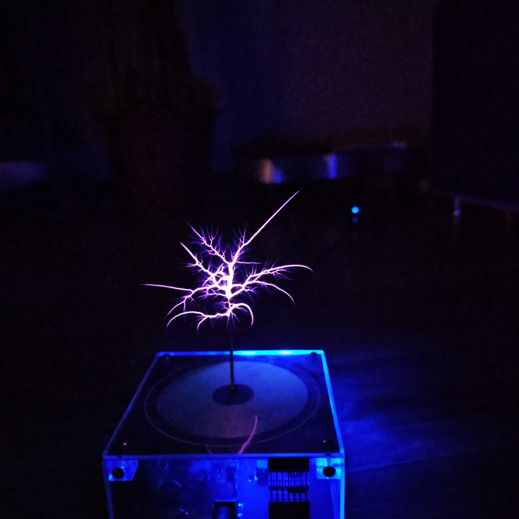 A photograph depicts a luminous Lichtenberg figure emanating from a device situated on a dark surface. The Lichtenberg figure, appearing as a branching, tree-like structure, is the focal point, radiating with a bright purple-white light. The device underneath appears to be an electrical apparatus housed in a clear rectangular casing, with illuminated blue edges and electronic components visible within. The background is dark, with some blurred shapes suggesting other objects in the room. There is no contextual information in the image to suggest that this image was taken in Rostov, Russia.