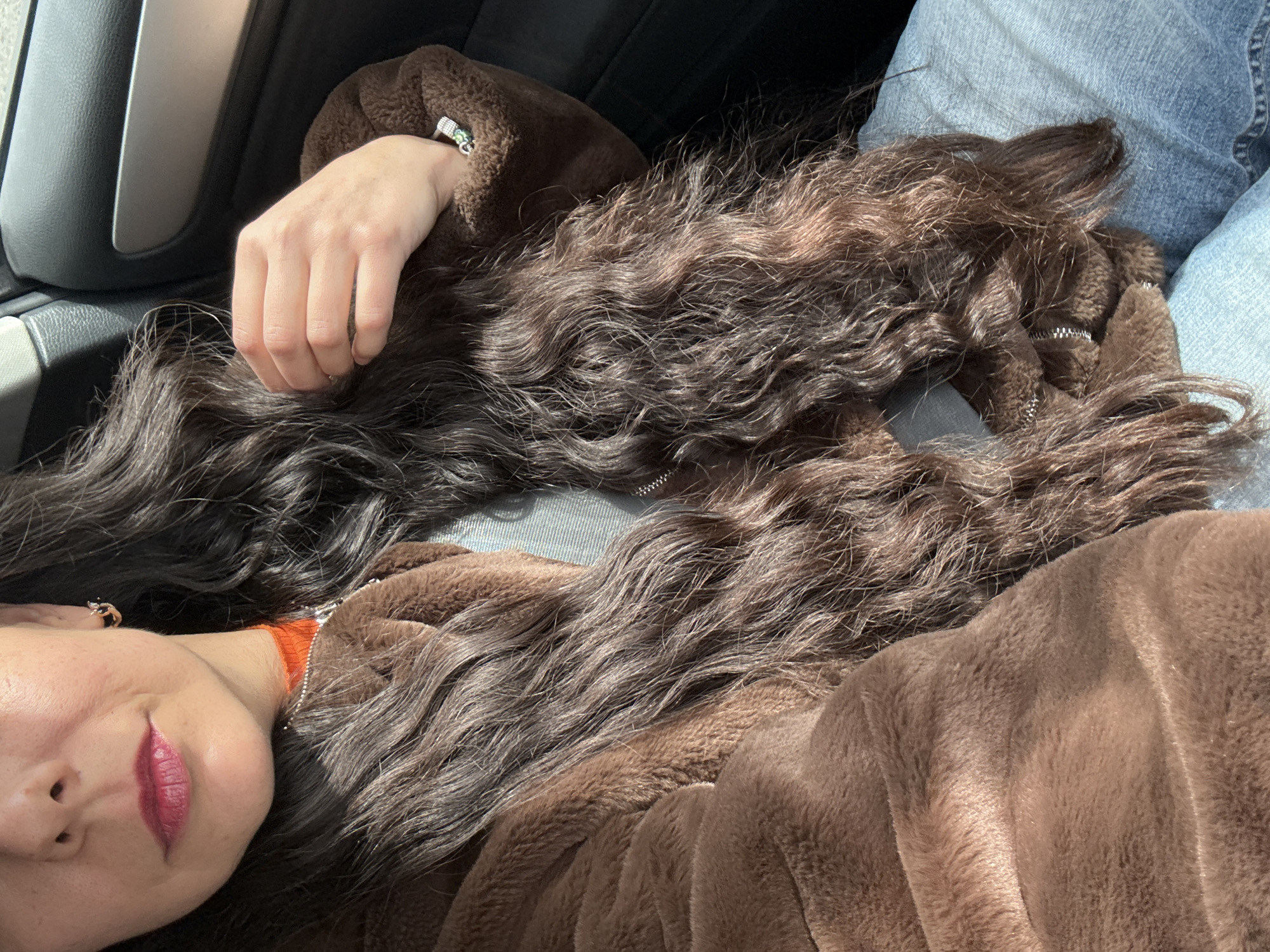 The image depicts a person lying down in what appears to be the passenger seat of a car. The person is wearing a brown, fleece-like jacket. They have long, dark brown hair that is spread out around them. Their face is partially visible, showing their lips with red lipstick. The person is wearing blue jeans. The interior of the car is visible in the background. The location is unknown and could not be determined based on the given picture.