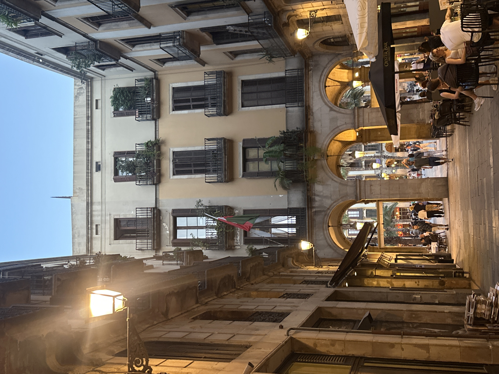 The image depicts a street scene in Barcelona, Spain, likely in the Gothic Quarter, characterized by narrow streets, traditional architecture, and outdoor dining areas. The perspective is looking down a cobblestone street lined with multi-story buildings on either side. The buildings feature stone facades, arched walkways on the lower levels, and balconies with wrought-iron railings on the upper floors. Many windows have wooden shutters, and some balconies have potted plants. A wrought-iron lamp is affixed to the left building, casting a warm glow.
In the mid-ground, arched openings lead to a wider area with palm trees visible. Outdoor tables and chairs are set up in front of establishments, with people seated at some of them. An umbrella with "OYSTER BAR" printed on it is visible above a dining area. Several people are walking along the street, some individually and some in small groups. The sky above is a clear, pale blue, suggesting it is late afternoon or early evening. The overall atmosphere is one of a European city street at dusk, with a lively but relaxed ambiance.