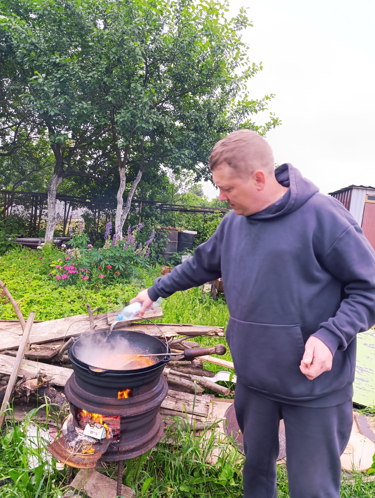 A medium- shot shows a Caucasian man in his late 30s, wearing a dark gray hooded sweatshirt and dark sweatpants, standing outside in a grassy area with trees and flowers in the background. He is holding a clear plastic bottle of water in his right hand and pouring liquid into a large black pot that is sitting on a makeshift stove. The stove appears to be made from car rims and is fueled by burning wood. Steam is rising from the pot, indicating that food is being cooked. The man is looking down at the pot. The background includes a wooden fence, some metal barrels, and a small building. The overall scene suggests an outdoor cooking activity.