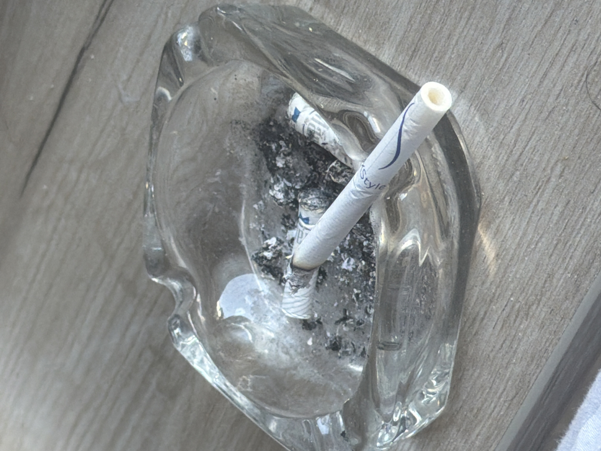 An overhead shot shows a clear glass ashtray containing cigarette butts and ash. One cigarette, branded "Style", is partially burnt and rests prominently in the ashtray. The ashtray sits on a wood-grain surface. There is no indication of the location being Podgorica, Montenegro. No people are present.