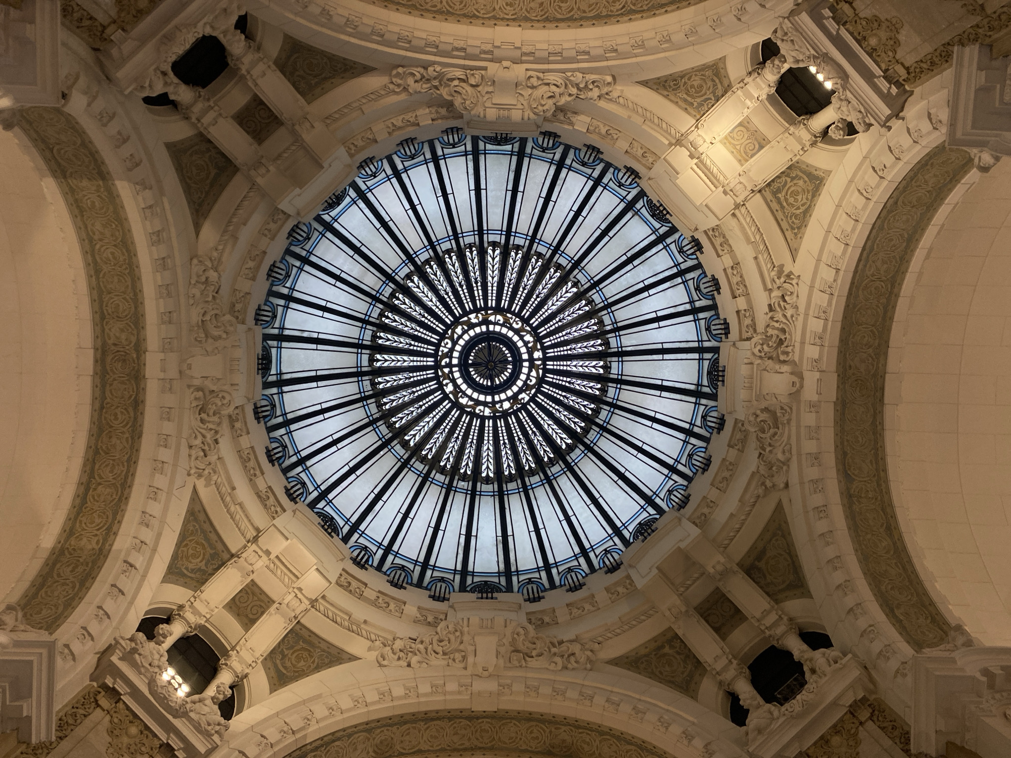 The image depicts the elaborate stained-glass dome of Galerías Pacífico, a shopping mall located in Buenos Aires, Argentina. The dome features a radial design with alternating clear and dark panels, surrounded by ornate architectural detailing, including arches, columns, and decorative moldings. The color palette is primarily white, beige, and gray with accents of black and blue in the glass dome.