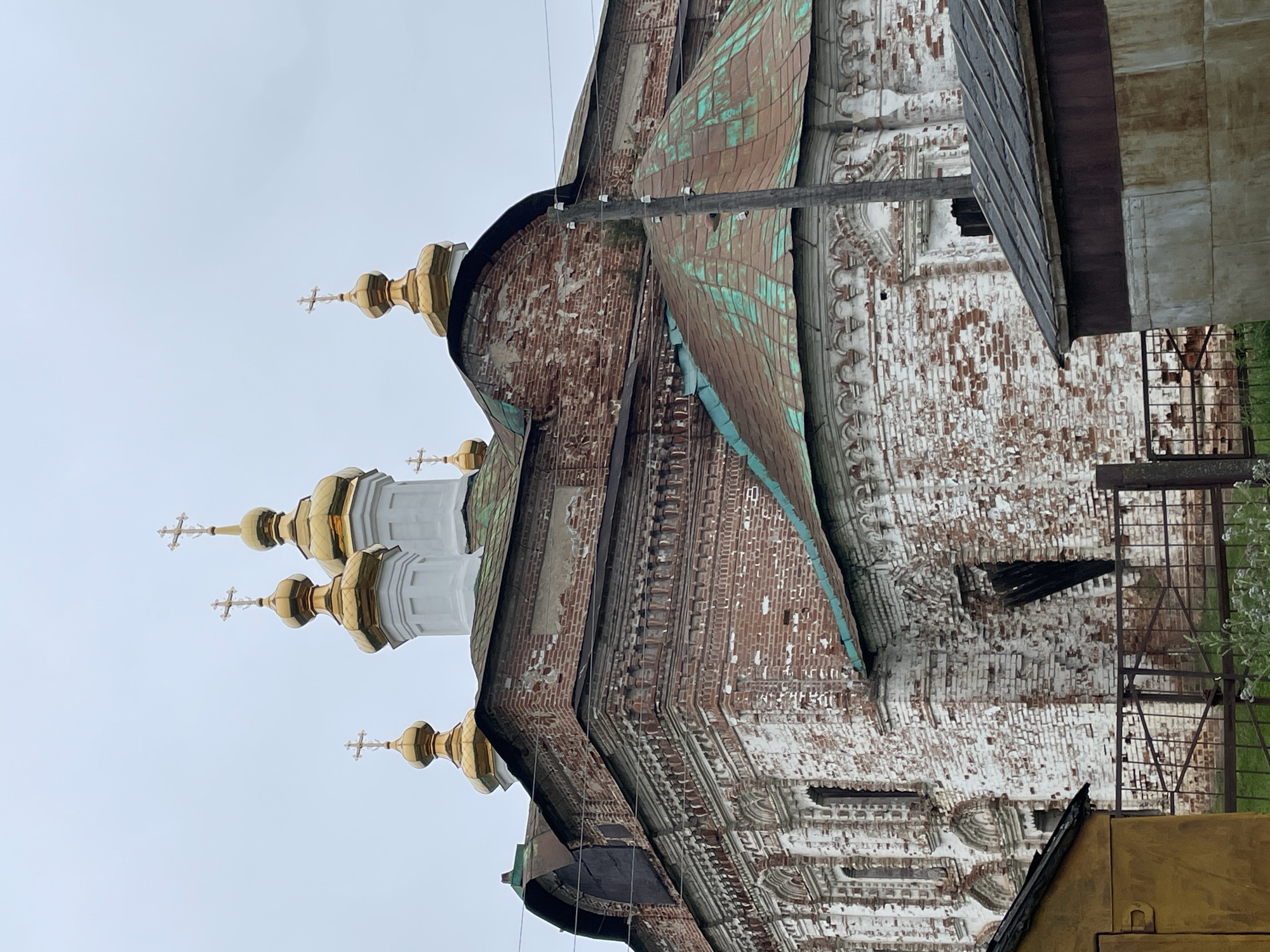 The image shows a close-up, low-angle view of the brick facade of an old, Orthodox church, likely located in Kstynino, Russia. The church is weathered, with areas of red brick exposed through peeling white paint. Several golden domes with crosses are visible at the top of the structure, indicating its religious purpose. One of the domes has a green, oxidized patina, suggesting age. The church has ornate architectural details, including decorative brickwork and arched window frames, many of which are dark and appear to be boarded up or empty. In the foreground, parts of simpler, more modern structures, including a yellow building and a corrugated metal shed, are visible. A metal fence runs across the lower part of the image. A utility pole with insulators stands to the right of the church. The sky is overcast, suggesting a cloudy day. No people are visible in the image.