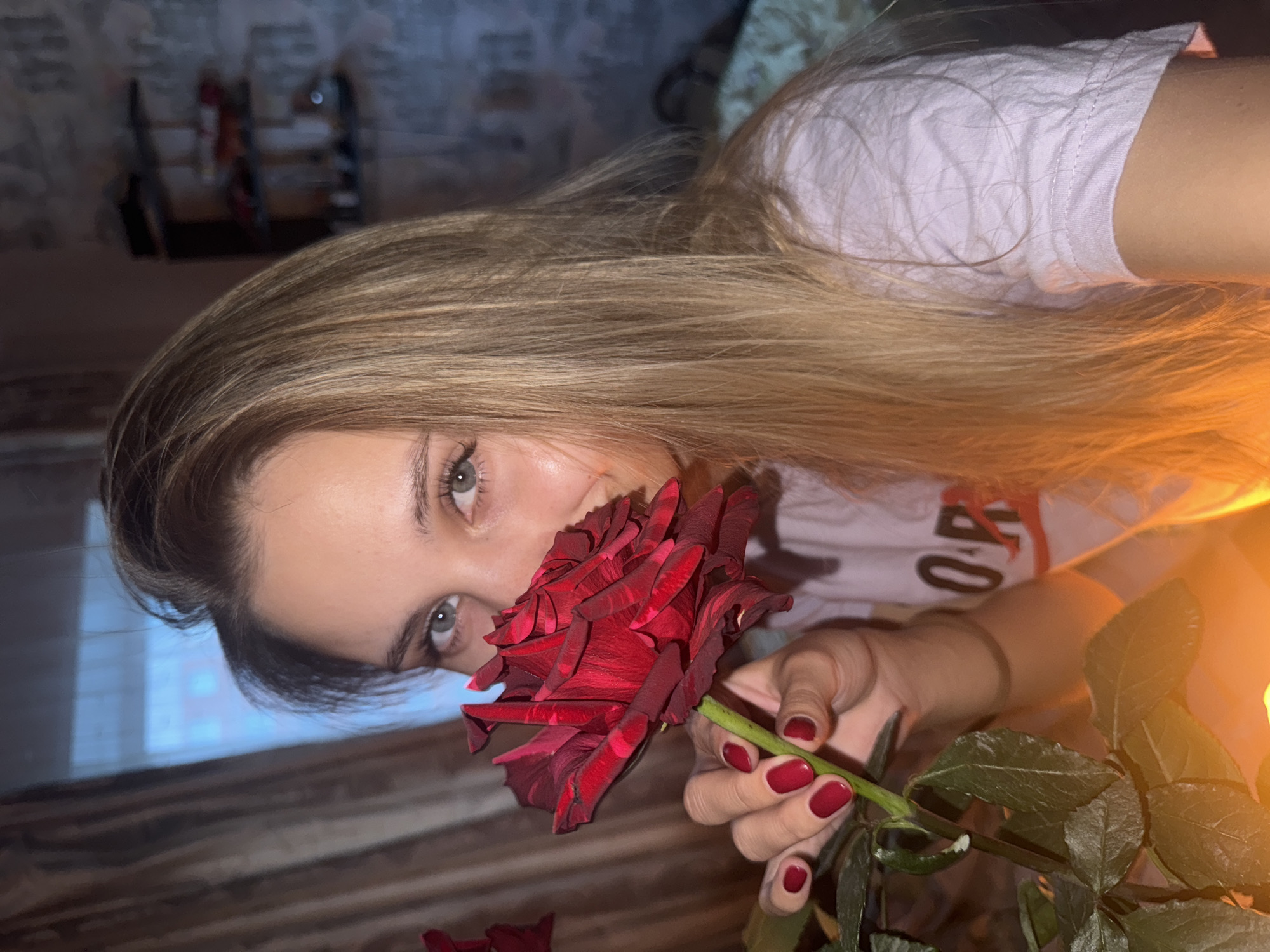 A young woman with long blonde hair and blue eyes is holding a dark red rose to her face, smelling it. She is wearing a light pink t-shirt with lettering on it. Her fingernails are painted a matching dark red. The background is blurred and appears to be indoors, with a window with curtains on the left and a shelf with objects on the right. There is a warm, orange glow emanating from the bottom of the image, possibly from a light source. The setting appears to be a domestic environment, potentially a bedroom or living room, in Chaikovsky, Russia.
