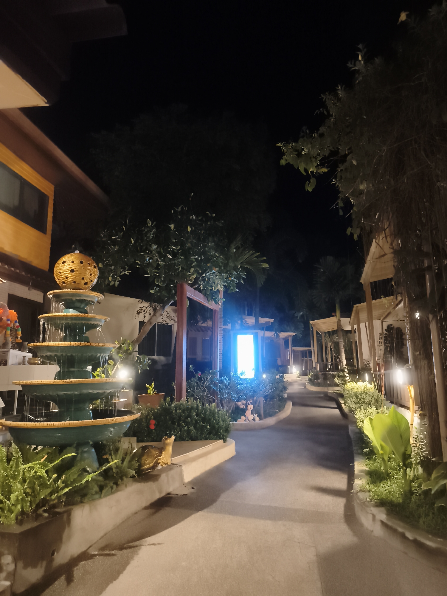 The image depicts an outdoor scene at night in Baan Chaweng, Thailand. A multi-tiered fountain with a decorative orb on top is in the foreground, to the left. The fountain is surrounded by greenery and a decorative golden turtle. A paved pathway illuminated by soft lighting leads into the distance between buildings. Lush tropical plants and small structures with wooden frames line the path, suggesting a resort or residential area. A bright blue light is visible further down the path. The sky is dark, indicating it's nighttime. No people are visible in the image.
