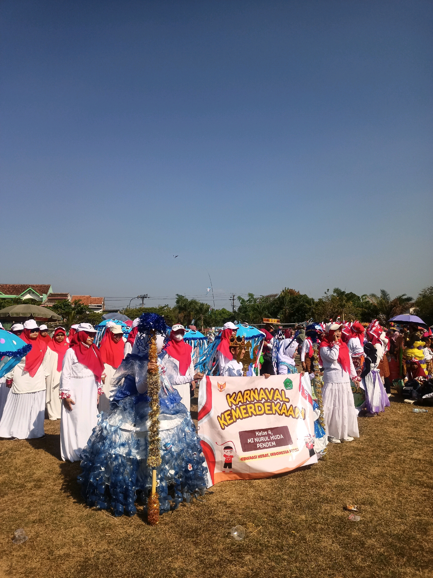 The image captures an outdoor event, likely a parade or festival, taking place on a grassy field under a clear blue sky. In the foreground, a group of people, mostly women and children, are dressed in colorful attire, suggesting a festive occasion. Several individuals are wearing white outfits with red headscarves, while others are adorned in elaborate costumes.
A prominent banner in the center of the image reads "KARNAVAL KEMERDEKAAN" (Independence Carnival), indicating the event's theme. Below this, smaller text states "Kelas 4, MI NURUL HUDA PENDEM" and "GENERASI HEBAT, INDONESIA KUAT" (Great Generation, Strong Indonesia). This suggests the participants are from a specific school or group.
One person in the foreground is wearing a striking blue and white costume, made from what appears to be recycled materials, possibly plastic bottles, with a shimmering gold garland. Other participants are holding decorative umbrellas, some in bright blue with patterns.
In the background, there are glimpses of houses with terracotta roofs and green trees, characteristic of a semi-urban or rural Indonesian setting. Power lines and poles are also visible. A few kites are flying in the distant sky. The ground is covered in dry grass, with some scattered litter, including a plastic bottle cap and some wrappers. The overall atmosphere appears to be bright and cheerful, with a sense of community participation. Based on the location information, this scene is likely in Sumberlawang, Indonesia.