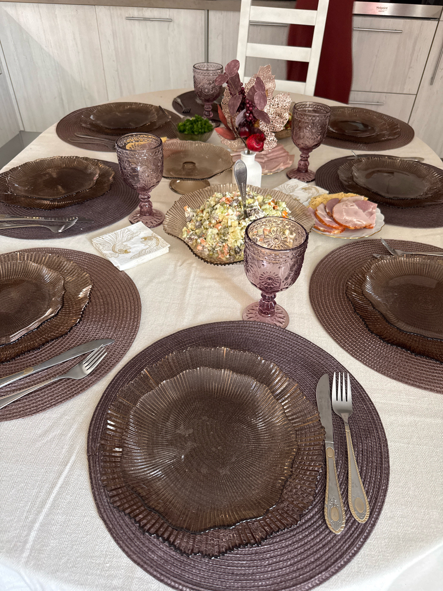 This is a close-up, high-angle shot of a dining table set for a meal. The table is covered with a light-colored tablecloth. On the table, there are several place settings, each with a dark, textured charger plate and a dark, patterned glass plate on top. Silverware, consisting of a knife and fork, is placed to the right of each setting. There are also several stemmed glasses with a decorative pattern in a mauve color. In the center of the table, a bowl of salad is visible, along with a small plate of sliced ham and some crackers. A decorative floral arrangement in autumnal colors is also present. The background shows a kitchen counter with cabinets and appliances. The overall impression is of a formal, yet possibly home-cooked, meal setting. The image does not provide any specific geographical information that would allow for its location to be determined within Saint Petersburg, Russia.
