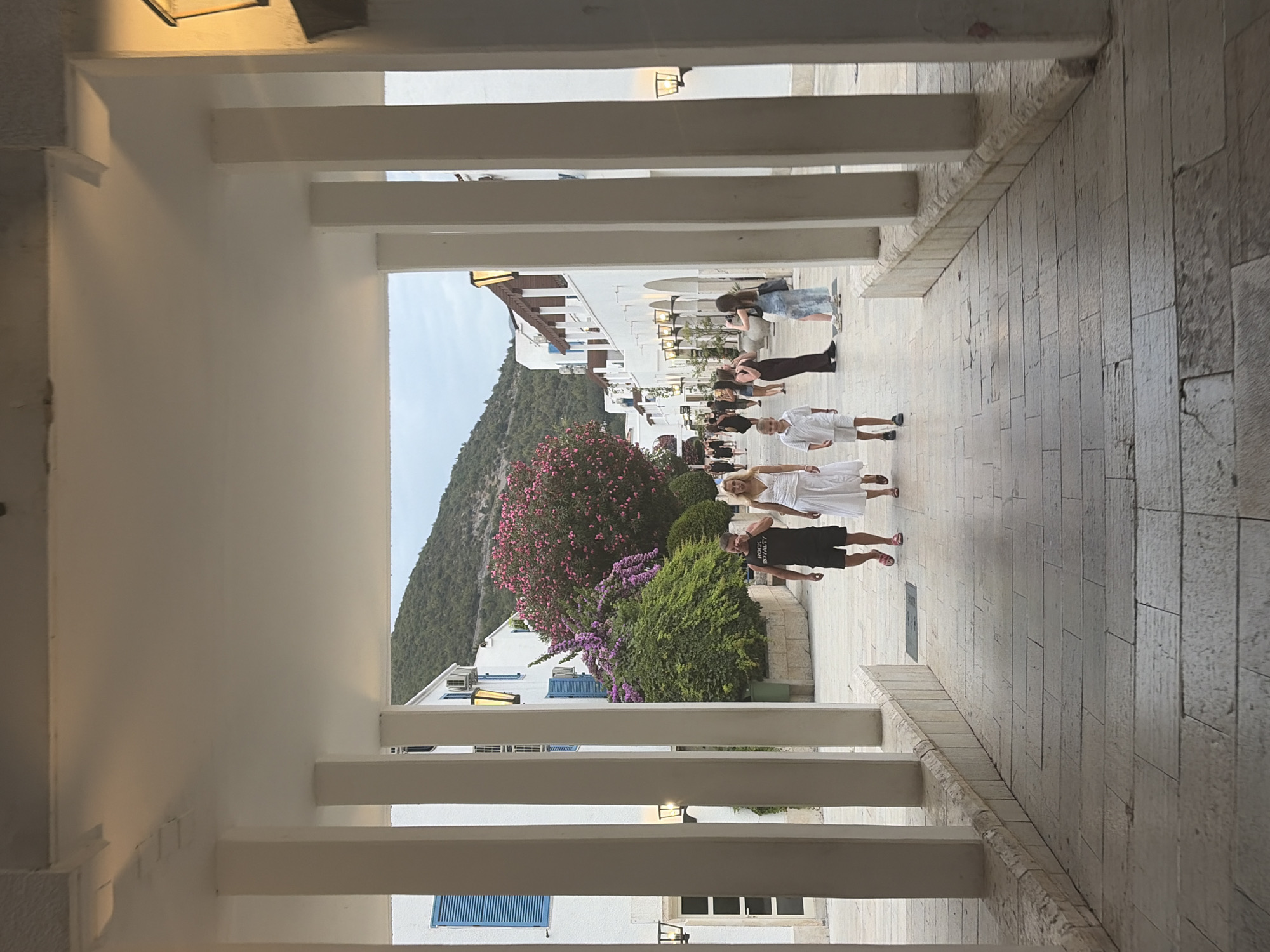 The image depicts a street scene in Budva, Montenegro, characterized by traditional Mediterranean architecture. White buildings with blue shutters line a paved walkway, suggesting a coastal town or resort. The foreground is dominated by a colonnade with large white pillars, framing a view of people walking along the street. A family, consisting of a man, woman, and two young boys, are walking towards the viewer. The man is wearing a black t-shirt and shorts, while the woman is in a white dress. One boy is in a white shirt and shorts, and the other is mostly out of frame. Other individuals are visible further down the street, engaged in various activities like walking and taking pictures. Lush green bushes and trees with vibrant pink flowers add a touch of nature to the urban environment. In the background, a verdant hillside rises, with the sky appearing overcast. The overall impression is that of a pleasant, leisurely stroll through a picturesque European town.