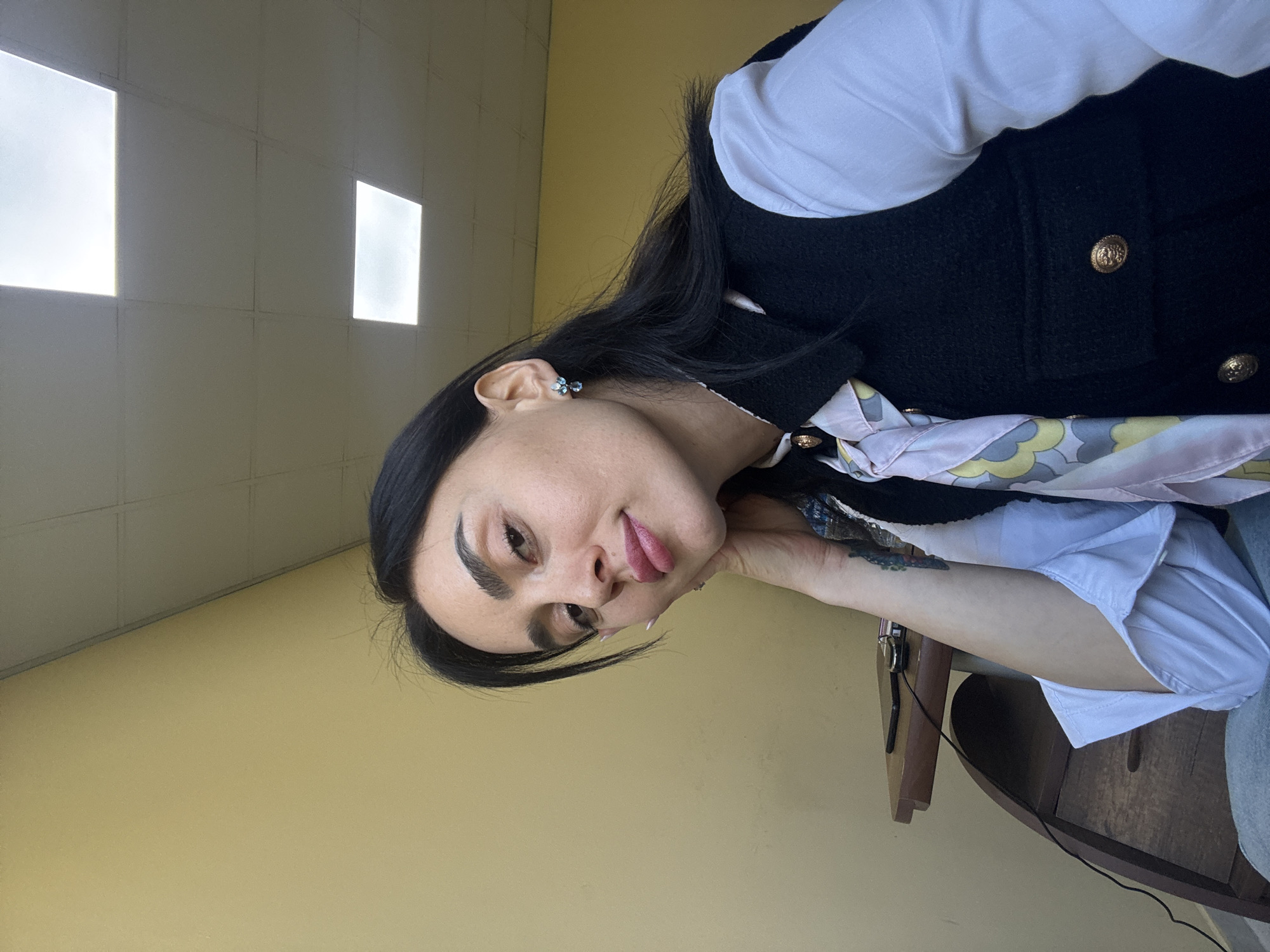 The image captures a woman in an indoor setting, likely posing for a photo. She appears to be in her 20s or 30s, with long, dark hair and dark, well-defined eyebrows. Her expression is playful, featuring a slight smile and her tongue peeking out. She looks directly at the camera, leaning her head on her left hand, which has a watch on the wrist and a partially visible tattoo on the forearm.
She is dressed in a white long-sleeved shirt, layered under a black textured sleeveless jacket adorned with gold buttons. A patterned scarf, featuring soft pink, yellow, and blue hues in an abstract design, is visible around her neck area. She wears small blue, flower-shaped earrings.
The background consists of a plain light yellow wall. Above, a light-colored ceiling with visible grid lines features two bright, rectangular recessed light panels, suggesting an institutional or office-like environment. A wooden table or desk is partially visible in the bottom right corner, with a black wire resting on it.
There are no specific visual cues to determine the time of day, but the artificial lighting is prominent. The overall mood is relaxed and casual. No text is visible in the image. The location context provided is Tbilisi, Georgia.