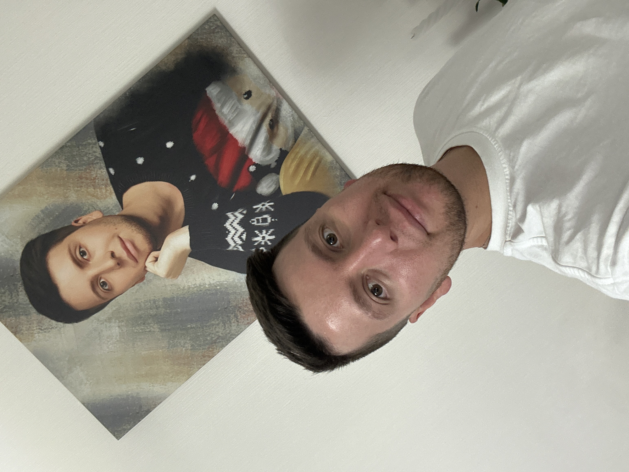A man in a white t-shirt stands in front of a wall displaying a painting of a man in a black sweater with a white pattern, a red hat, and holding a white object, likely a toy or stuffed animal. The location is possibly in the city of Мирный, Russia.