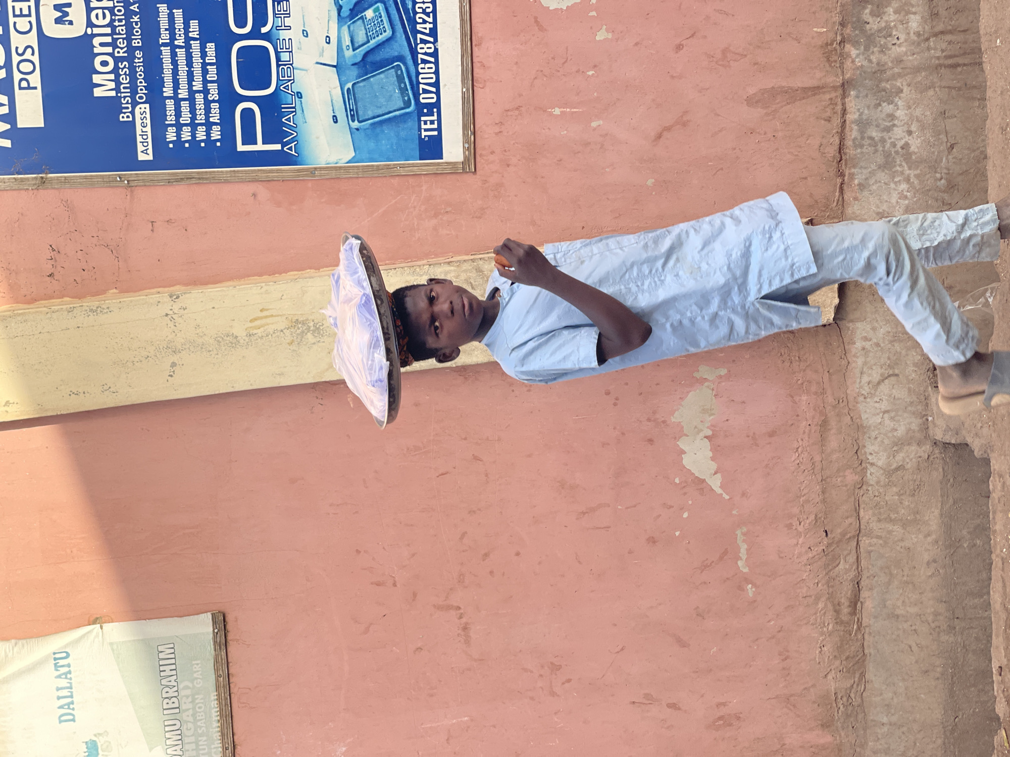 The image, taken in Samaru, Nigeria, features a young male standing outdoors against a reddish-pink wall. He is positioned centrally, looking directly at the camera with a neutral expression. He wears a light blue long-sleeved shirt and matching trousers. On his head, he balances a flat, circular tray covered with a translucent white or light purple plastic bag, suggesting he is carrying goods for sale or transport. In his right hand, he holds a small, peeled orange fruit, possibly an orange or tangerine.
The background consists primarily of a faded, somewhat rough reddish-pink wall that shows signs of age and wear. Above the boy, a blue sign with white text is affixed to the wall. This sign advertises "Monier Business Relation" and services such as "Issue Moniepoint Terminal," "Open Moniepoint Account," "Issue Moniepoint Atm," and "Sell Out Data." A phone number, "TEL: 0706787423," is also visible. Below the boy and to the left, a white and green sign is partially visible, showing text like "DALLATU" and "AMU IBRAHIM HIGARI TUN SABON GARI Chairman." The ground beneath him is uneven, appearing to be a mixture of dirt and concrete. The bright, uniform lighting indicates it is daytime, capturing a moment of daily life in an urban or semi-urban setting.