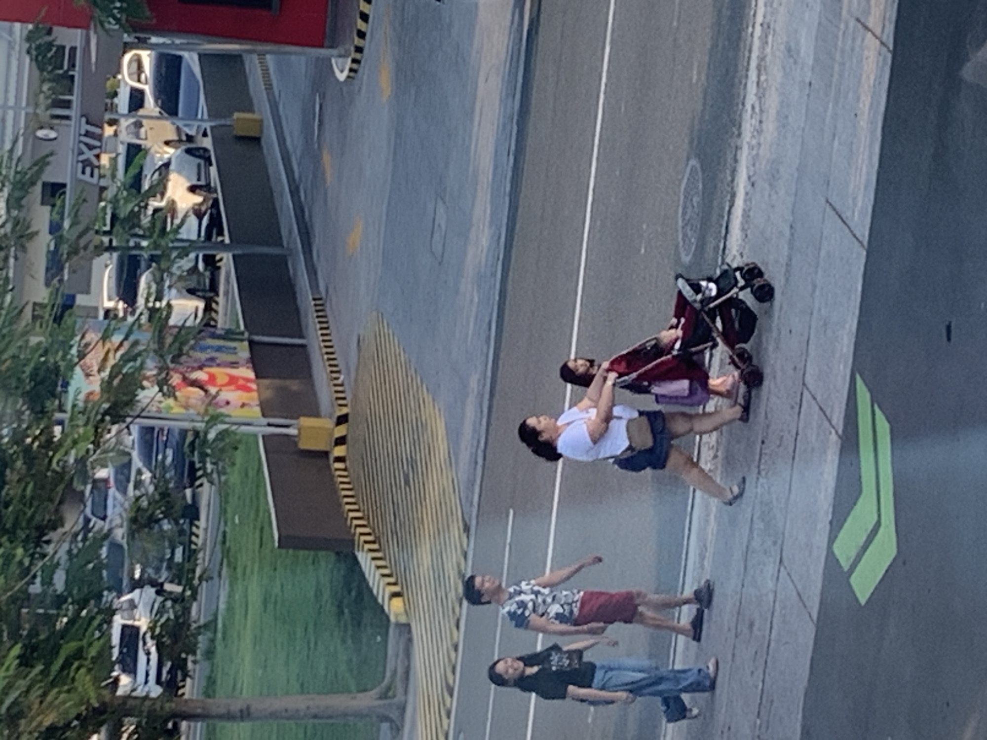 In Angeles City, Philippines, a low-angle medium shot captures a sidewalk and road scene. On the sidewalk, four individuals are visible. From left to right, a young woman in a black t-shirt and blue jeans walks with a slight smile. Next to her, a man in a camouflage t-shirt and red shorts walks, also with a subtle smile. Further to the right, a woman wearing a white t-shirt and dark shorts pushes a stroller containing a young child dressed in red. The stroller is dark-colored with red accents. Behind them, another child is partially visible in the stroller. The group appears to be walking together on the sidewalk adjacent to a paved road. In the background, cars are parked in a lot, and an "EXIT" sign is prominent. Trees and greenery are scattered throughout the scene, and a brightly colored mural is visible on a building in the distance. Yellow and black striped barriers are present along the edge of the road and sidewalk. A green arrow painted on the road indicates a direction. The overall lighting suggests it is daytime.