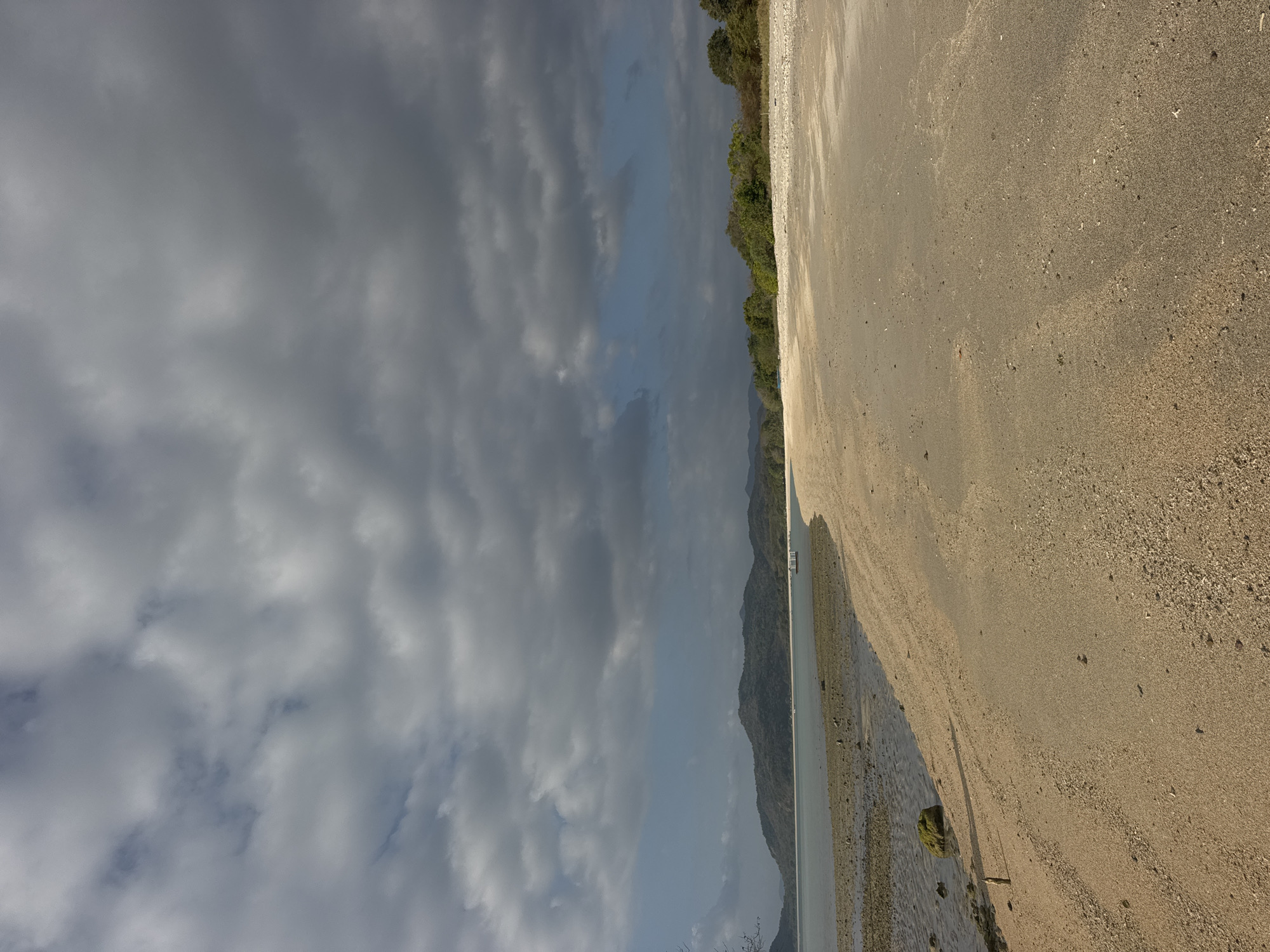A wide shot captures a tranquil beach scene under a dramatic, cloudy sky. The foreground is dominated by a sandy beach, with subtle textures of sand and small pebbles. To the left, the water gently laps the shore, revealing a rocky and mossy seabed exposed at low tide. A solitary piece of driftwood stands upright in the sand. In the middle ground, the calm ocean stretches towards a distant, tree-covered mountain range. A small, white floating structure is visible in the water near the shoreline. The sky above is a canvas of dark, brooding clouds, with patches of bright blue sky peeking through, suggesting the potential for changing weather. The scene evokes a sense of peaceful isolation and natural beauty.