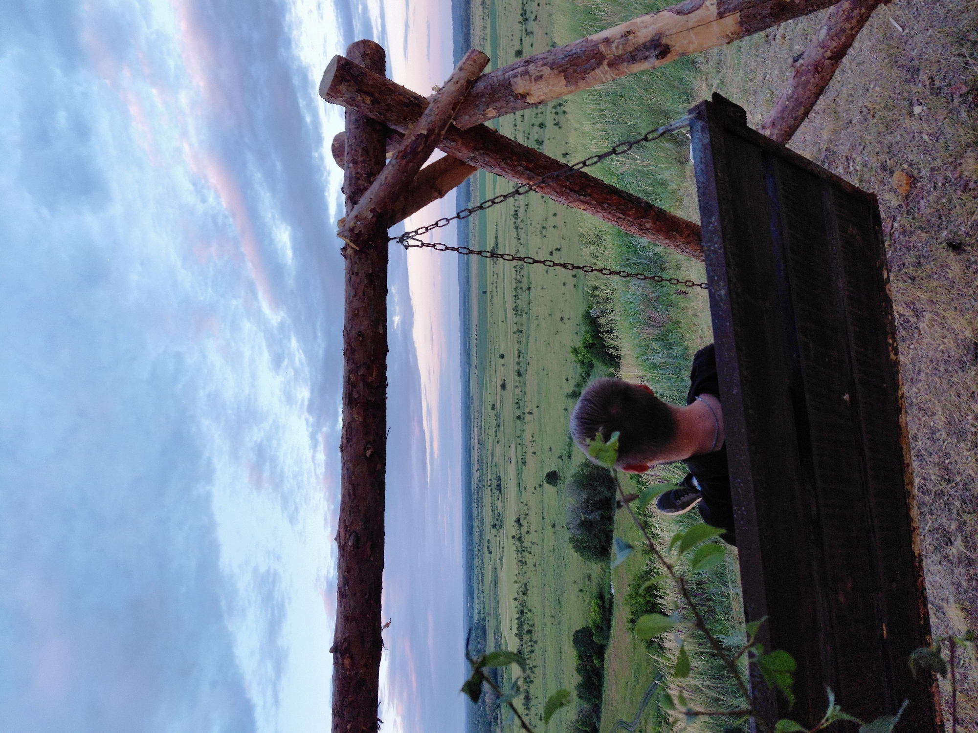 An eye-level, outdoor shot captures a person on a wooden swing overlooking a vast, green landscape. The sky is cloudy and appears to be twilight. The swing is made of dark wood with a rough texture, suspended by metal chains from a rustic wooden frame constructed from logs. The person is facing away from the camera, seated on the swing, wearing a black shirt and dark pants, with their legs crossed. In the foreground, leafy branches partially obscure the view. The background reveals a sprawling field with scattered trees and undulations, hinting at a rural or natural setting. The overall impression is one of peaceful contemplation amidst nature.